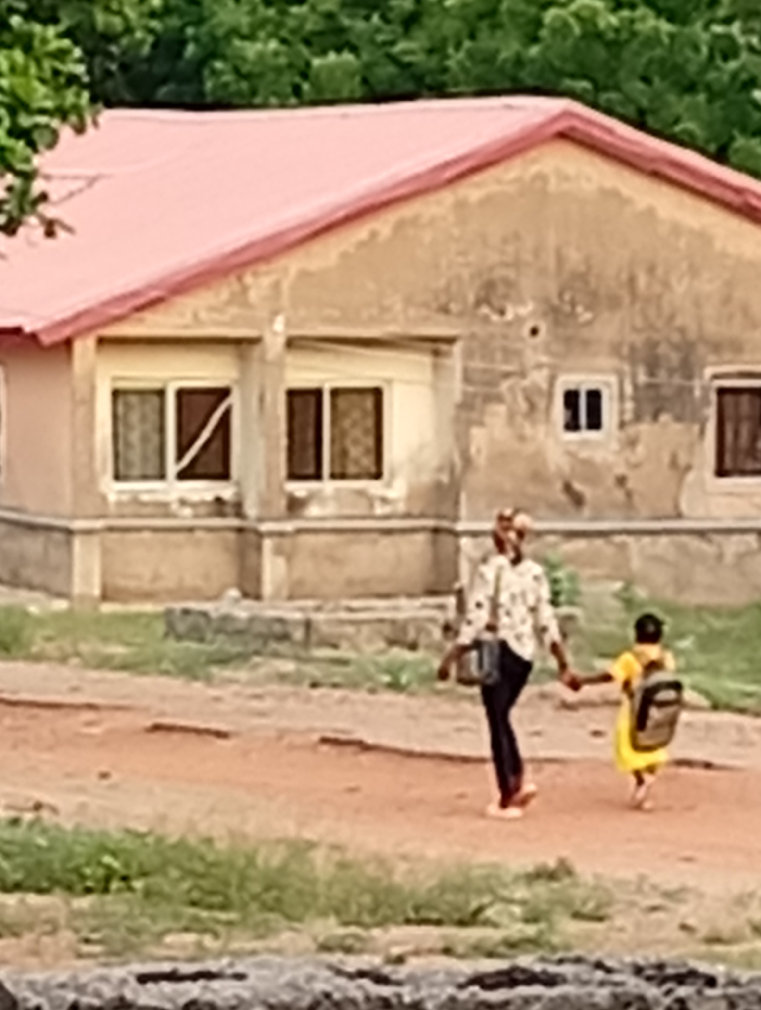 In Adankolo, Nigeria, a person and a child are walking hand-in-hand on a dirt road in front of a single-story building. The building has a red roof and beige walls with several windows. The child wears a yellow dress and carries a backpack. The adult wears a light-colored patterned shirt and dark pants, carrying a bag on their shoulder. Greenery is visible in the background and foreground. The image has a slightly blurred quality.