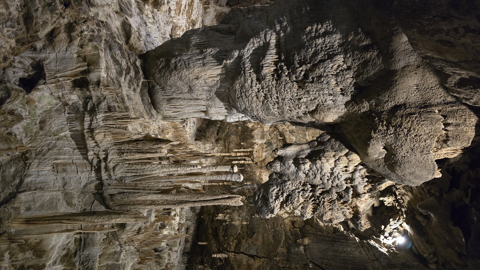 This is an image of the interior of a cave, likely in the area of Vavřinec, Czechia. The cave is filled with various speleothems, which are mineral deposits formed in caves over long periods. In the foreground, there are large stalagmites and stalactites. Stalagmites are formations that grow upward from the cave floor, while stalactites hang down from the ceiling. The formations are primarily composed of limestone, giving them a textured, rocky appearance.
The lighting in the cave is artificial, casting shadows that highlight the intricate details and rough surfaces of the speleothems. The colors are predominantly earthy tones of brown, gray, and beige, with variations in shade depending on the lighting and mineral composition. The overall environment is dark and damp, typical of a natural underground cave system. There are no people or animals visible in the image. The focus is entirely on the geological formations and the natural architecture of the cave. The background shows more of the cave walls and ceiling, also adorned with smaller stalactites and rock formations.