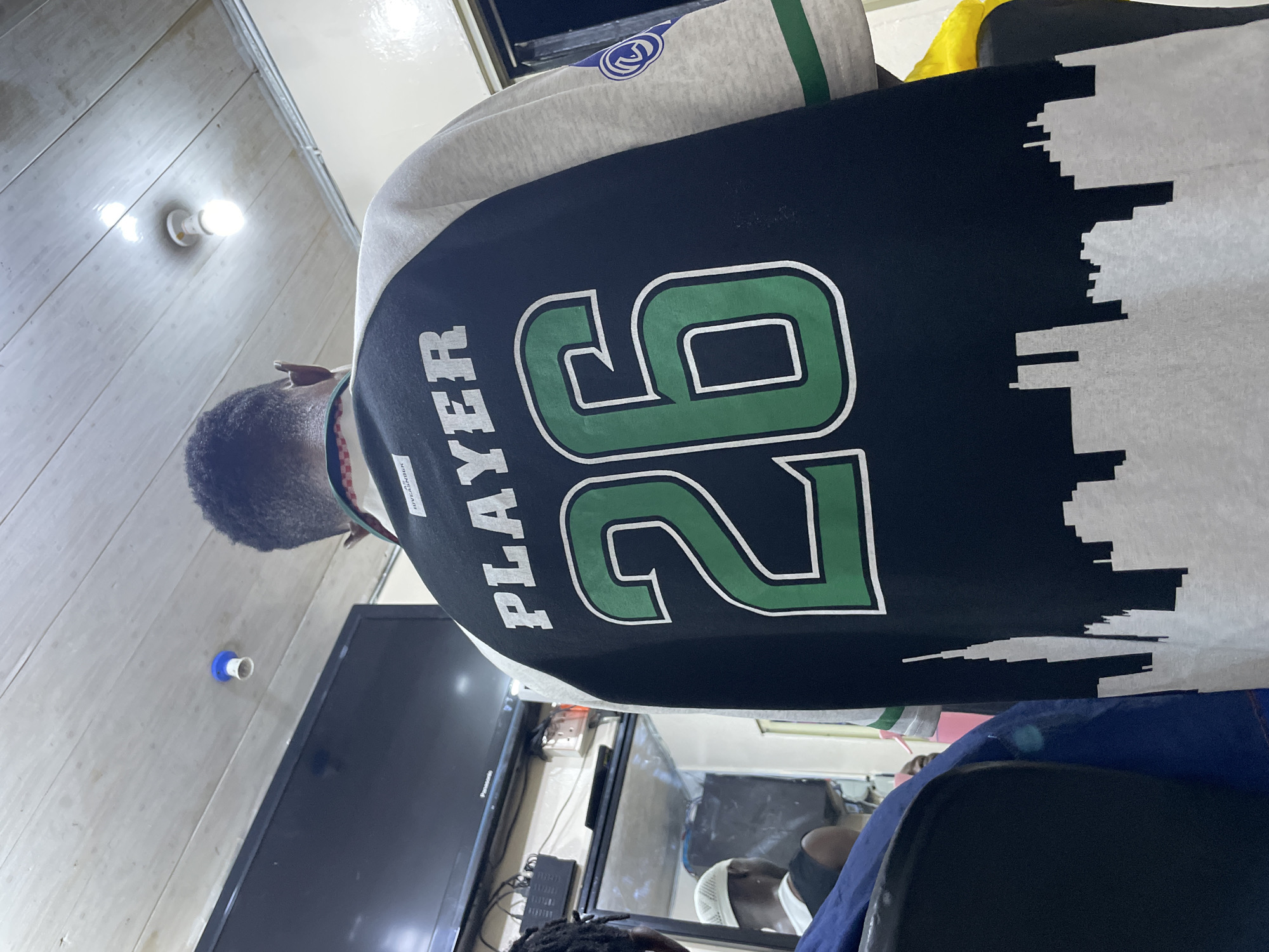 A person wearing a jacket with the word "PLAYER" and the number "26" on the back is indoors. The jacket is black with white and green accents, and a white skyline design is on the right side. A television is visible in the background. The location may be Ado, Nigeria.