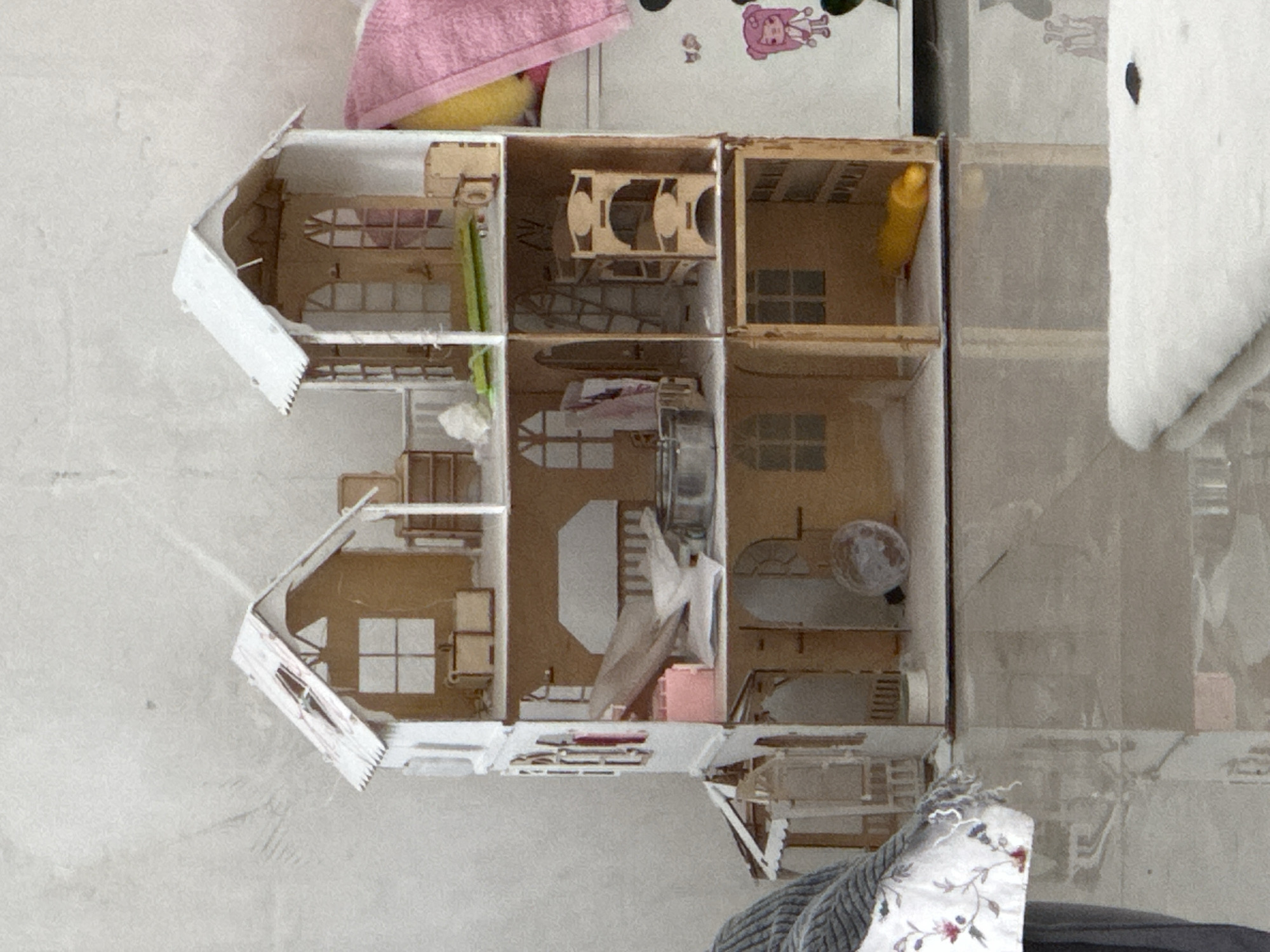 A high-angle shot shows a partially constructed dollhouse sitting on a surface that appears to be a floor or tabletop. The dollhouse has two sections, one above the other, with multiple rooms in each section. The rooms are in various states of assembly, some with furniture and other miniature items inside. The structure is made of a light-colored material, possibly wood or cardboard, and parts of it are painted white. The surrounding environment includes other objects such as a pink towel, a white plush toy, a patterned pillow, and what looks like wall or cupboard in the background. The location is unclear, with no definitive markers to indicate a specific place.