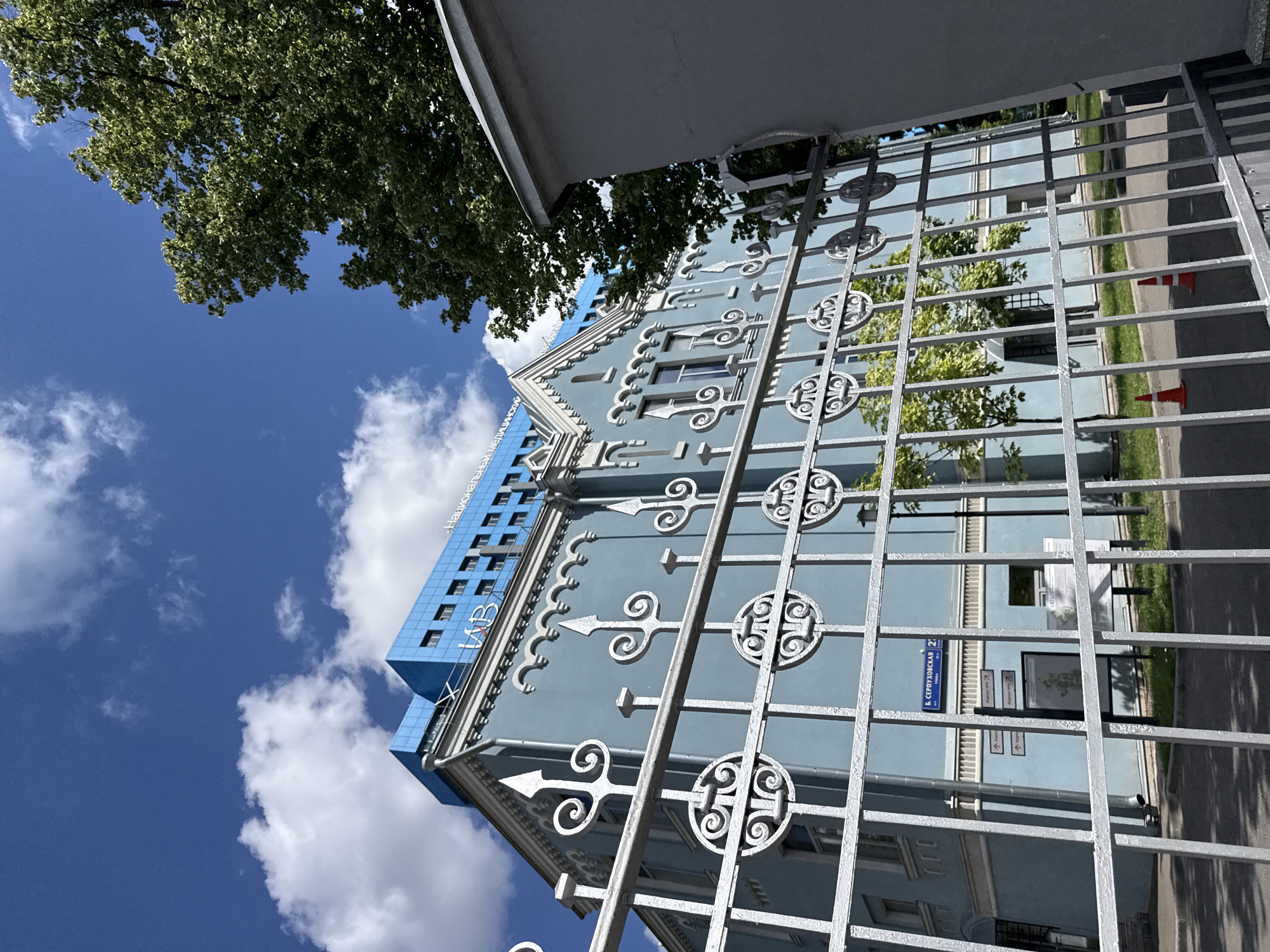 The image shows a pale blue building with an ornate white fence in the foreground. The building has multiple windows and a decorative roofline. On top of the pale blue building is a modern blue structure with text that reads "НАЦИОНАЛЬНЫЙ МЕДИЦИНСКИЙ ЦЕНТР". Through the fence, a street sign is visible that reads "Б. СЕРПУХОВСКАЯ 27". This indicates the location is Bolshaya Serpukhovskaya Street, building 27, in Moscow, Russia. Two orange traffic cones are on the asphalt in the middle ground. A large green tree is to the right of the blue structure. The sky above is blue with scattered white clouds.