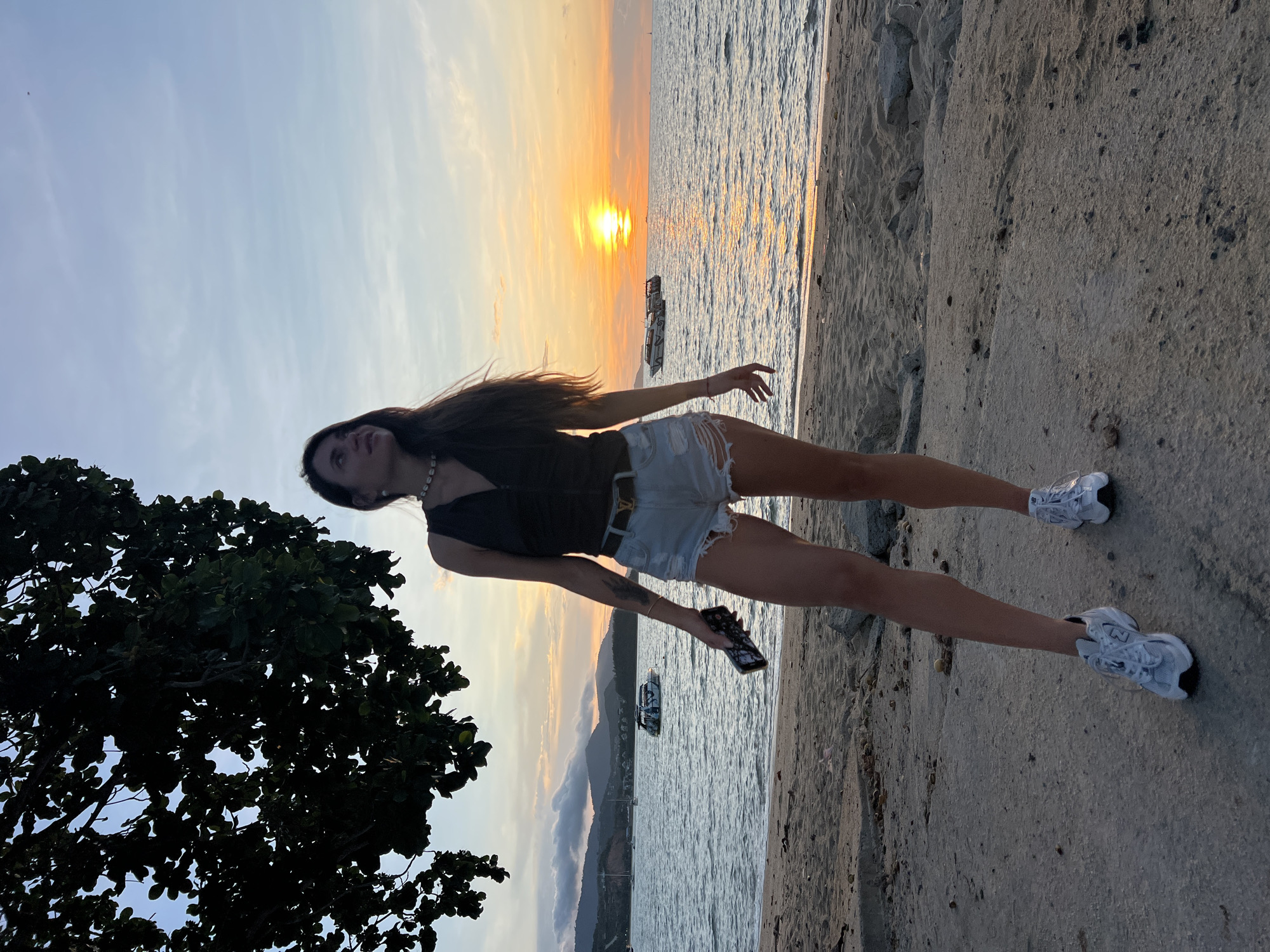 A young woman stands on a sandy beach in Baan Bang Rak, Thailand, during sunset. She is looking up and to her right. She has long, dark hair and is wearing a black tank top, ripped denim shorts, and white sneakers. She is holding a black smartphone in her right hand. The sea is calm with a few boats in the distance. The sky is a gradient of orange, yellow, and blue. A large tree with dark green leaves is in the upper left corner of the image.