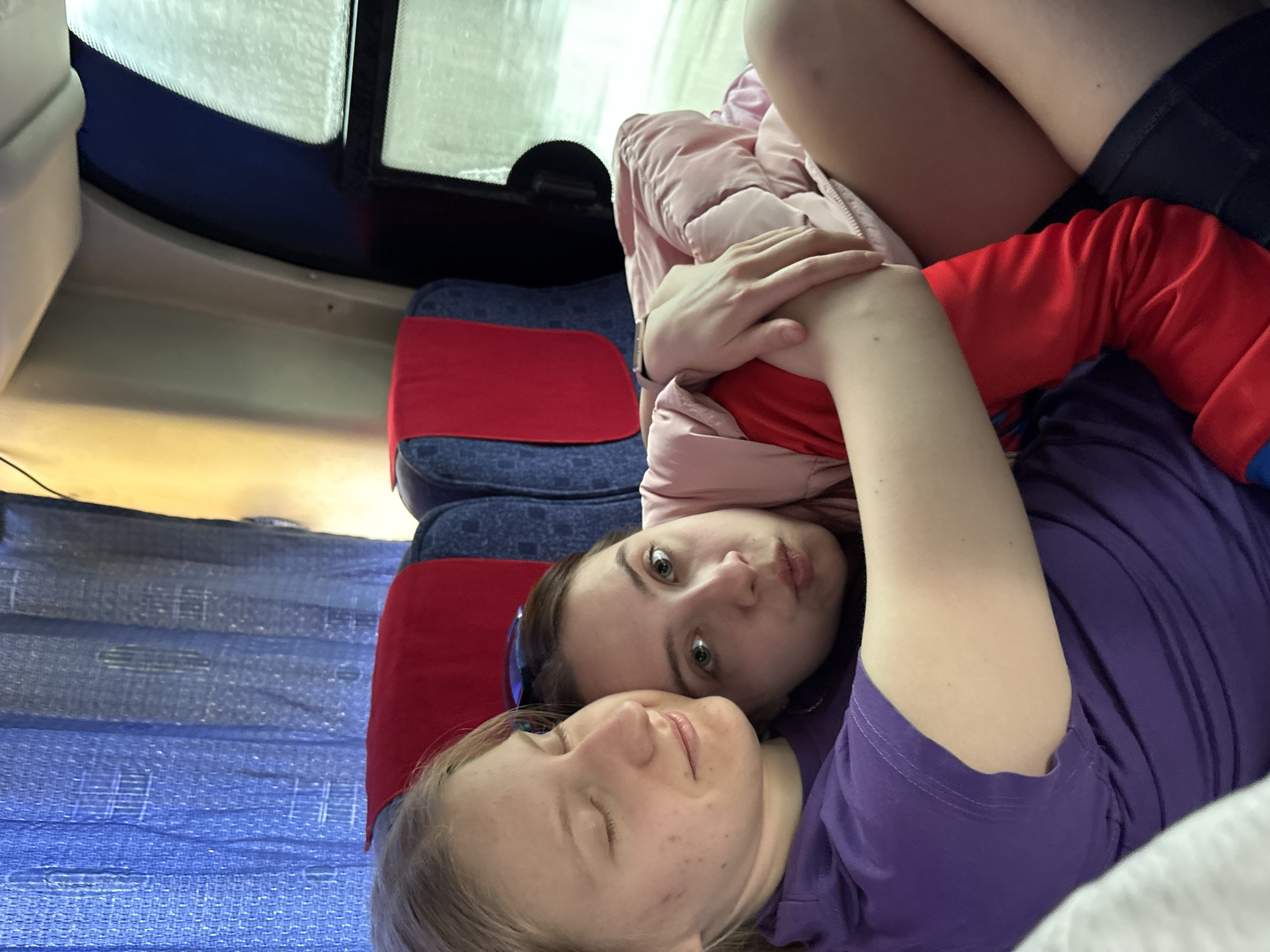 Two young women are closely positioned together. One woman, with blonde hair and closed eyes, is wearing a purple shirt. The other woman, with brown hair and open eyes, is wearing a pink jacket over a red shirt and black shorts. They are seated on a blue and red patterned seat, possibly on a bus or train. A window is visible in the upper left corner of the image. The location is Verkhnyaya Dobryanka, Russia.