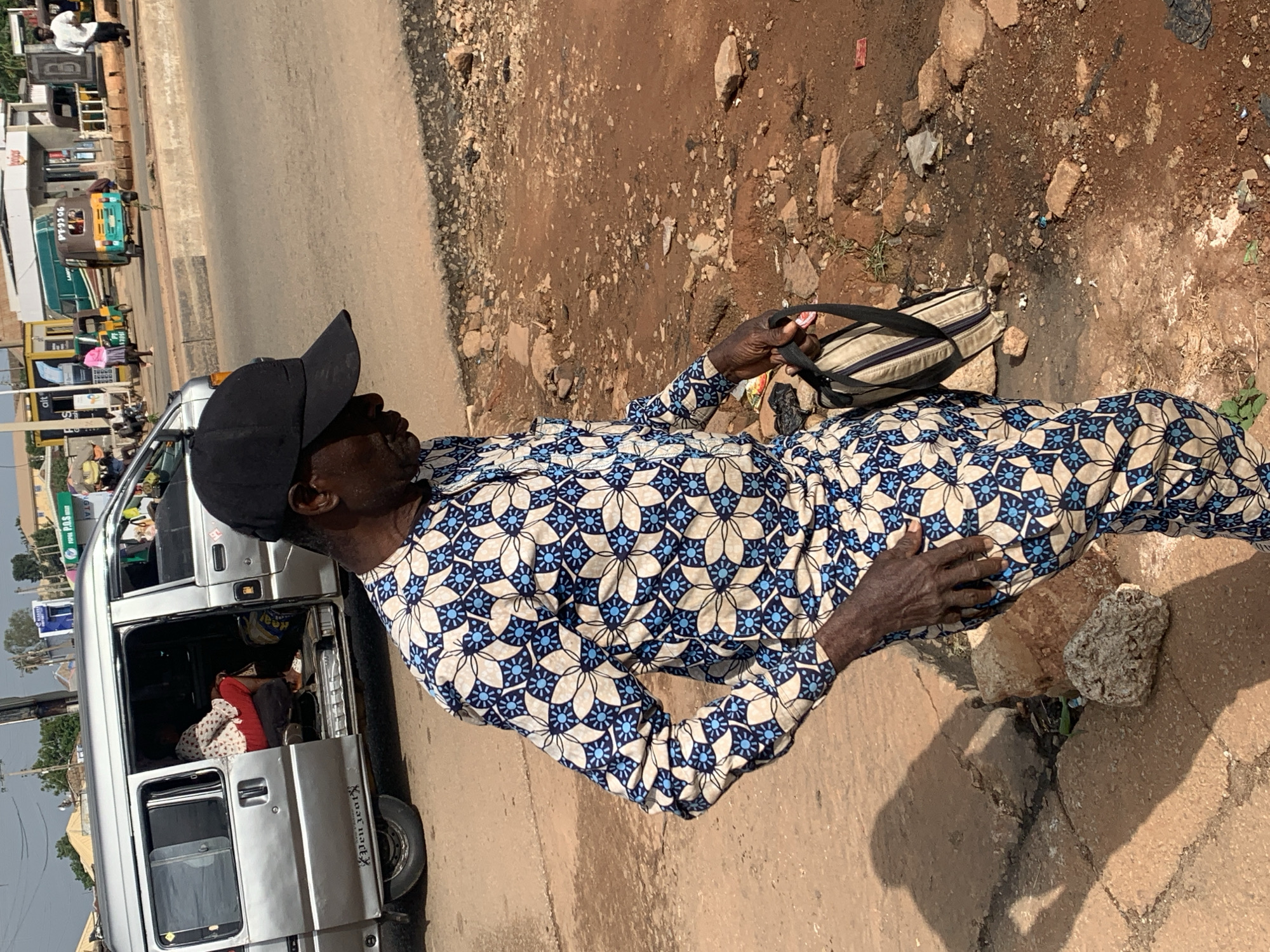 In Gura Top, Nigeria, a man wearing a black baseball cap and a blue and white patterned outfit stands on a dusty, reddish-brown street next to a silver minivan. The minivan's side door is open, revealing a woman sitting inside. The man is holding a light-colored bag with black straps in his right hand and has his left hand on his hip. The street is littered with rocks and debris. In the background, other vehicles, including a green bus and a yellow auto-rickshaw, are visible on a paved road, along with buildings and trees, suggesting an urban or semi-urban environment.