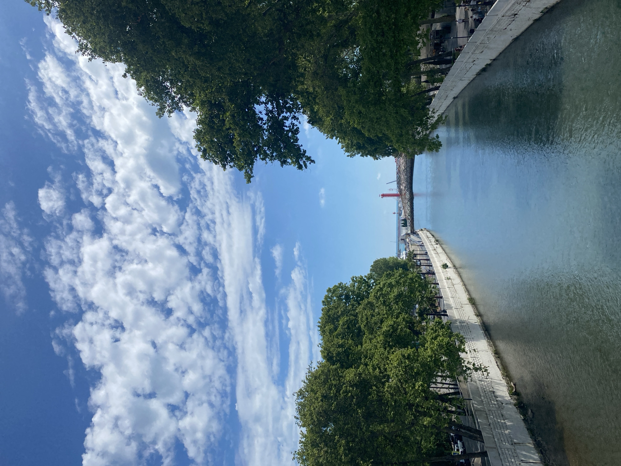This is a medium-angle, full shot of a wide canal in Sochi, Russia, on a clear, bright day with scattered clouds. The canal runs from the foreground to the background, where it opens to the sea. The water is a deep blue-green, reflecting the sky and trees. On either side of the canal are wide, paved promenades lined with mature trees and low, white, concrete walls. The left promenade is lined with parked cars and buildings in the distance. The right promenade has pedestrians walking and buildings with awnings. In the far distance, a red lighthouse is visible near the sea, along with a modern, curved bridge spanning the canal. The sky is a vibrant blue, with fluffy white clouds creating shadows on the water. The overall scene depicts a scenic, urban waterfront environment.