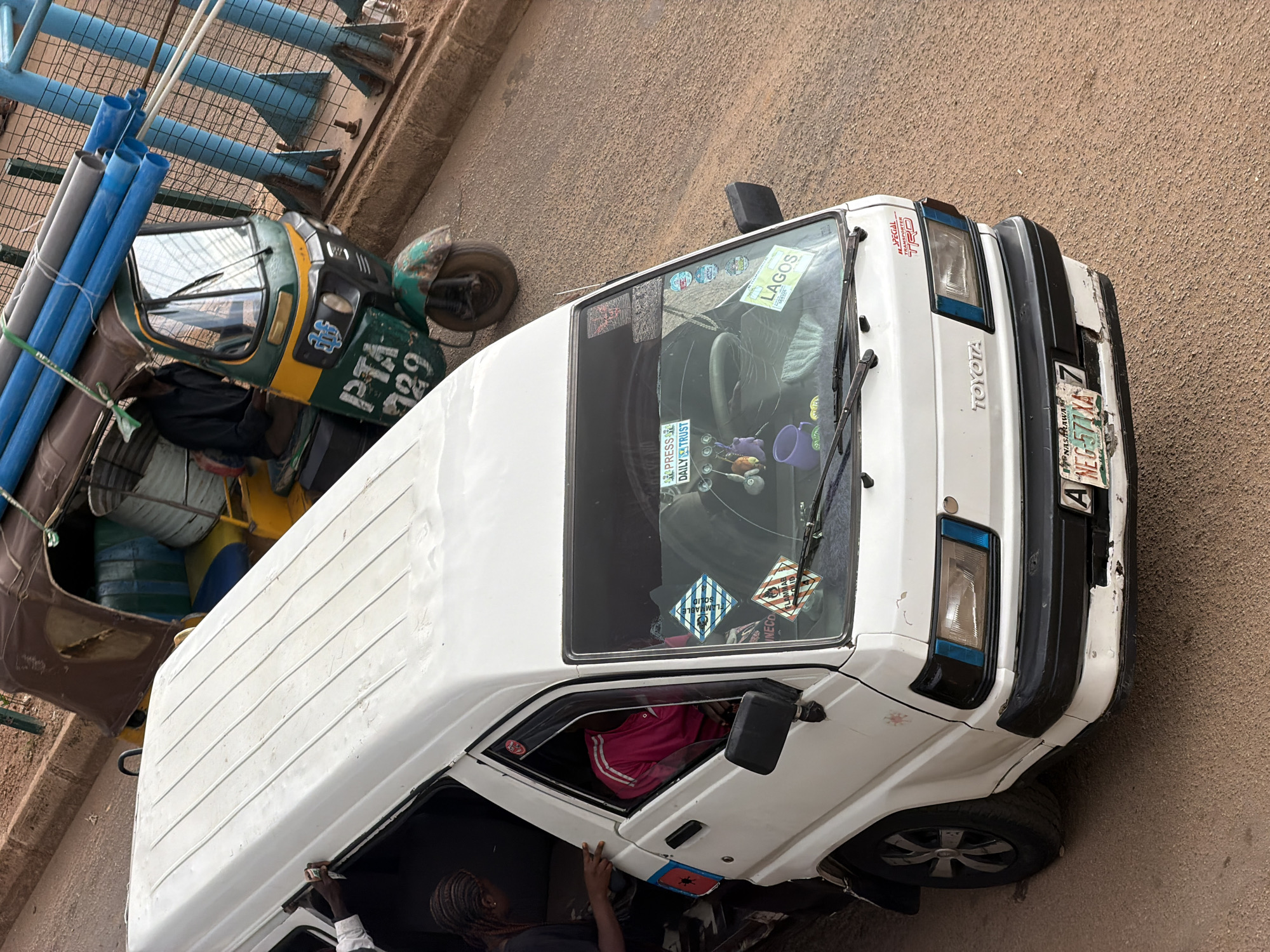 In Bukuru, Nigeria, a white Toyota minibus is parked on a dirt road. The minibus has several stickers on its windshield, including one that says "PRESS DAILY TRUST" and another that says "LAGOS". A person wearing a pink shirt is visible inside the minibus. In the background, a green and yellow auto-rickshaw is loaded with blue and grey pipes. The rickshaw has "PTA 729" written on its front. The environment appears to be a dusty, outdoor area with a fence in the background.