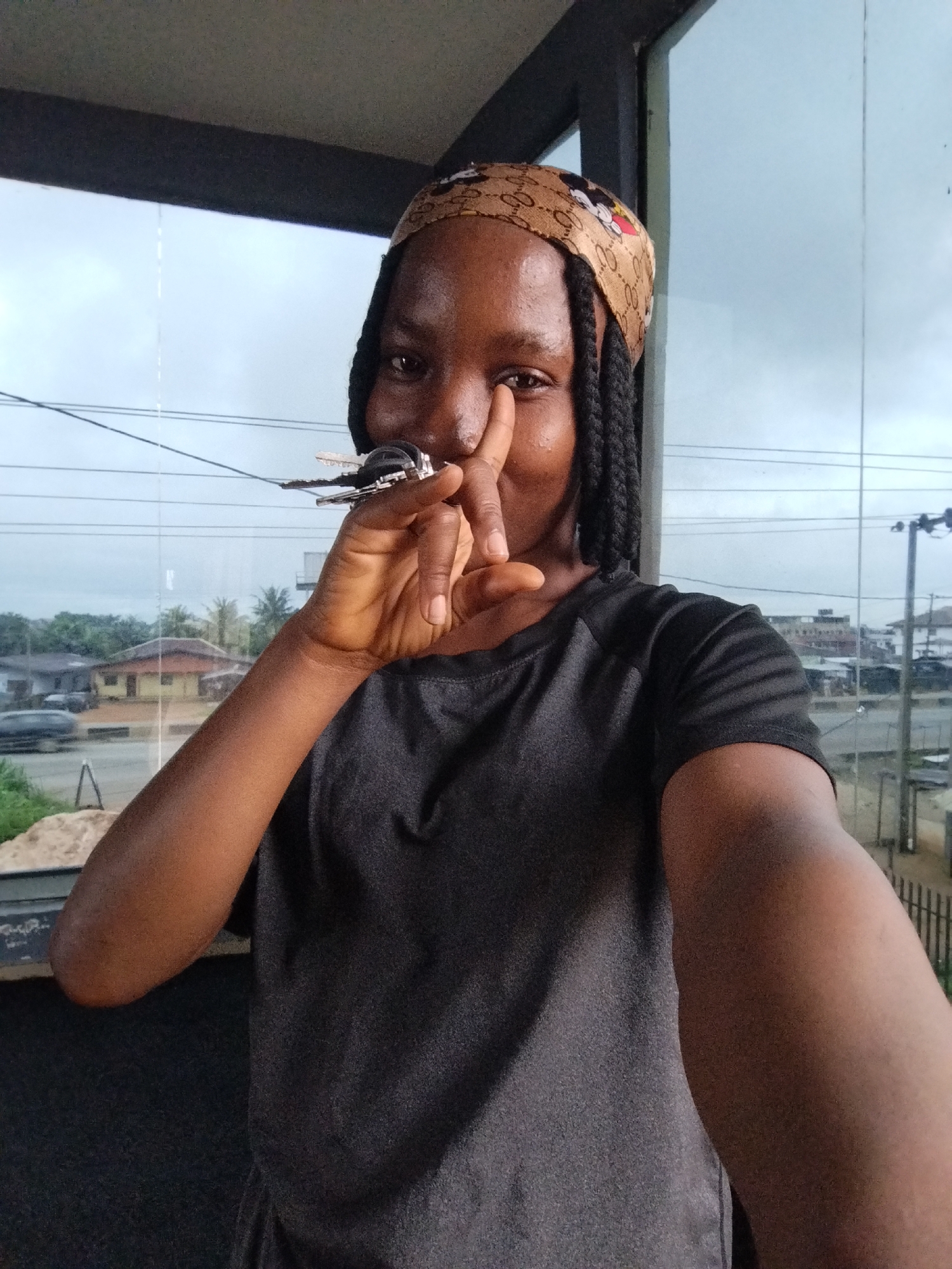 A person with dark skin and braided hair wears a bandana patterned with Mickey Mouse and the Gucci logo, along with a dark gray t-shirt. They are holding a set of keys to their nose. The background is a blurry urban landscape, with buildings, trees, and power lines visible through a large window. The lighting suggests an overcast day.