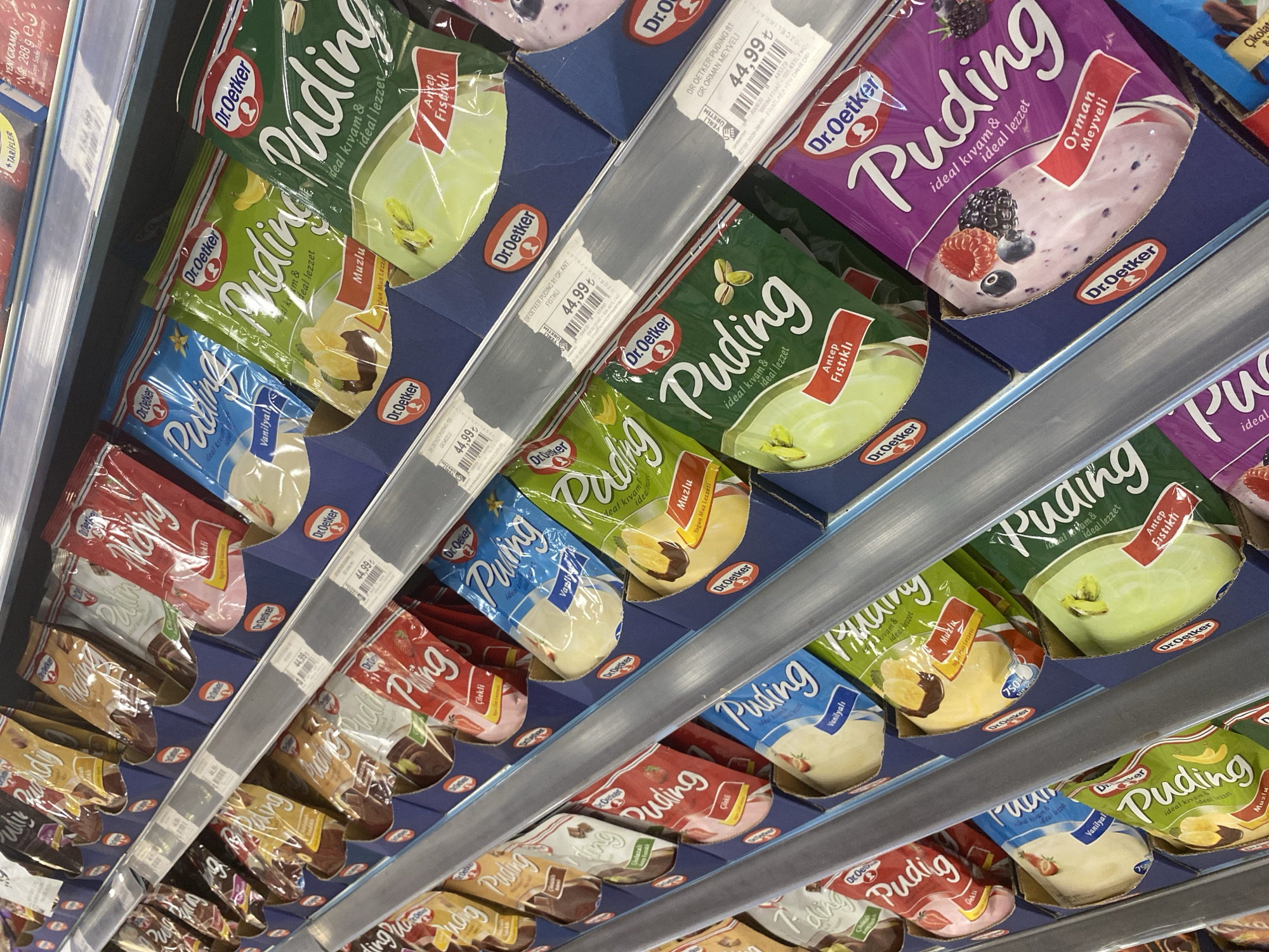 The image shows a retail display of Dr. Oetker pudding products on shelves in a supermarket. The products are packaged in colorful sachets with the Dr. Oetker logo and pudding flavors like Vanilla, Banana, Pistachio, and Forest Fruit. Some of the sachets have prices displayed on them, such as 44.99 ₺. The display is organized by flavor and arranged in cardboard holders. The overall lighting suggests an indoor commercial environment. The scene is consistent with a grocery store in Konya, Turkey, given the currency and likely language of the product labeling. There are no people visible in the image.