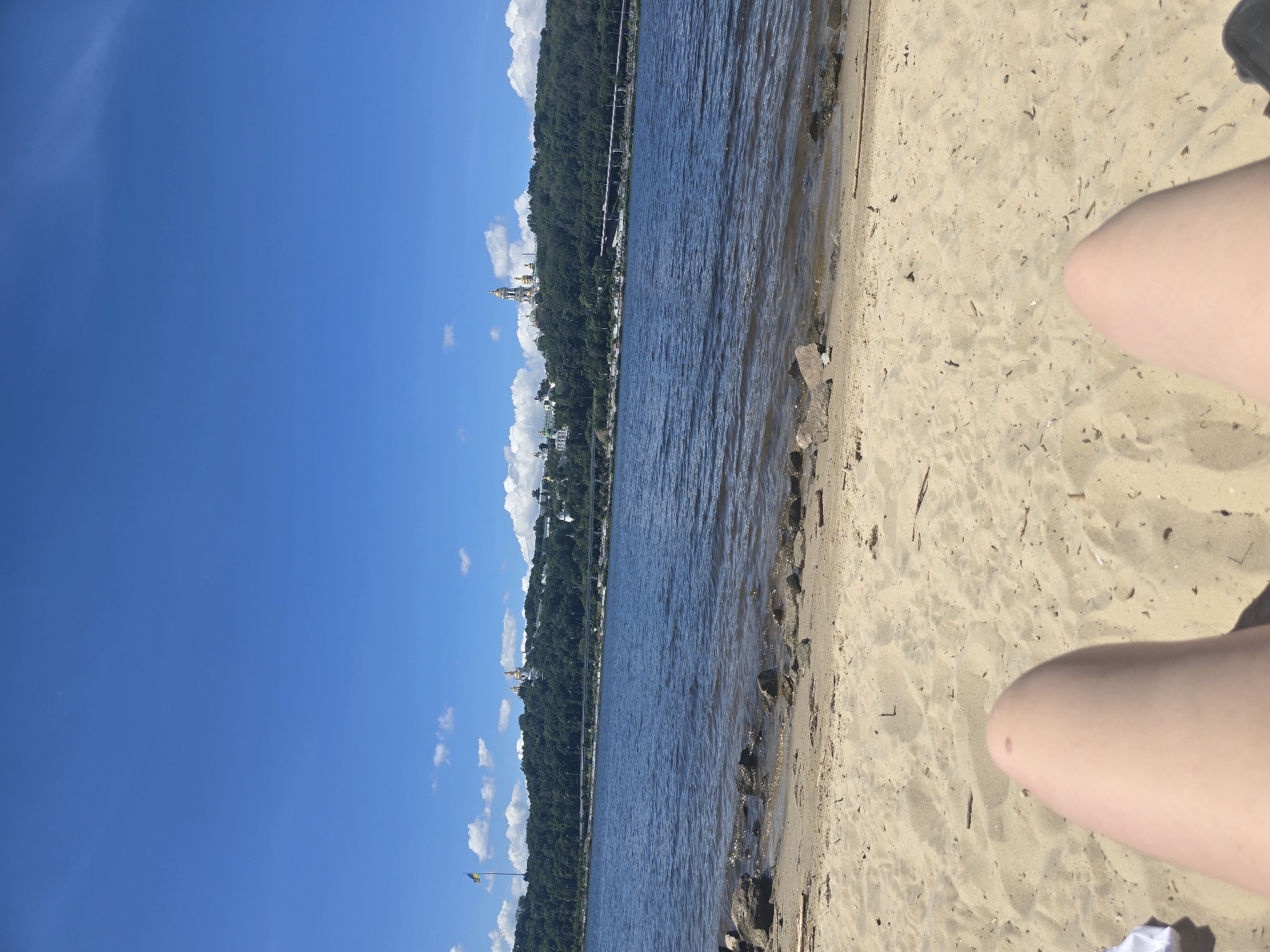 A first-person perspective shot captures a sandy beach on a sunny day, with the viewer's legs visible in the foreground, relaxed on the sand. Beyond the beach, a body of water stretches out, with gentle ripples on its surface. In the distance, a verdant hillside rises, densely covered with trees. Atop the hill, several white buildings with golden domes, characteristic of Orthodox churches, are prominent. To the left of the churches, a tall flagpole flies the blue and yellow flag of Ukraine. A multi-lane bridge crosses the water in the middle ground, connecting the two sides of the city. The sky above is a vibrant, clear blue, dotted with a few scattered white clouds. This scene is indicative of the area near the Kyiv Pechersk Lavra, a historic Orthodox monastery complex located on the right bank of the Dnieper River in Kyiv, Ukraine.