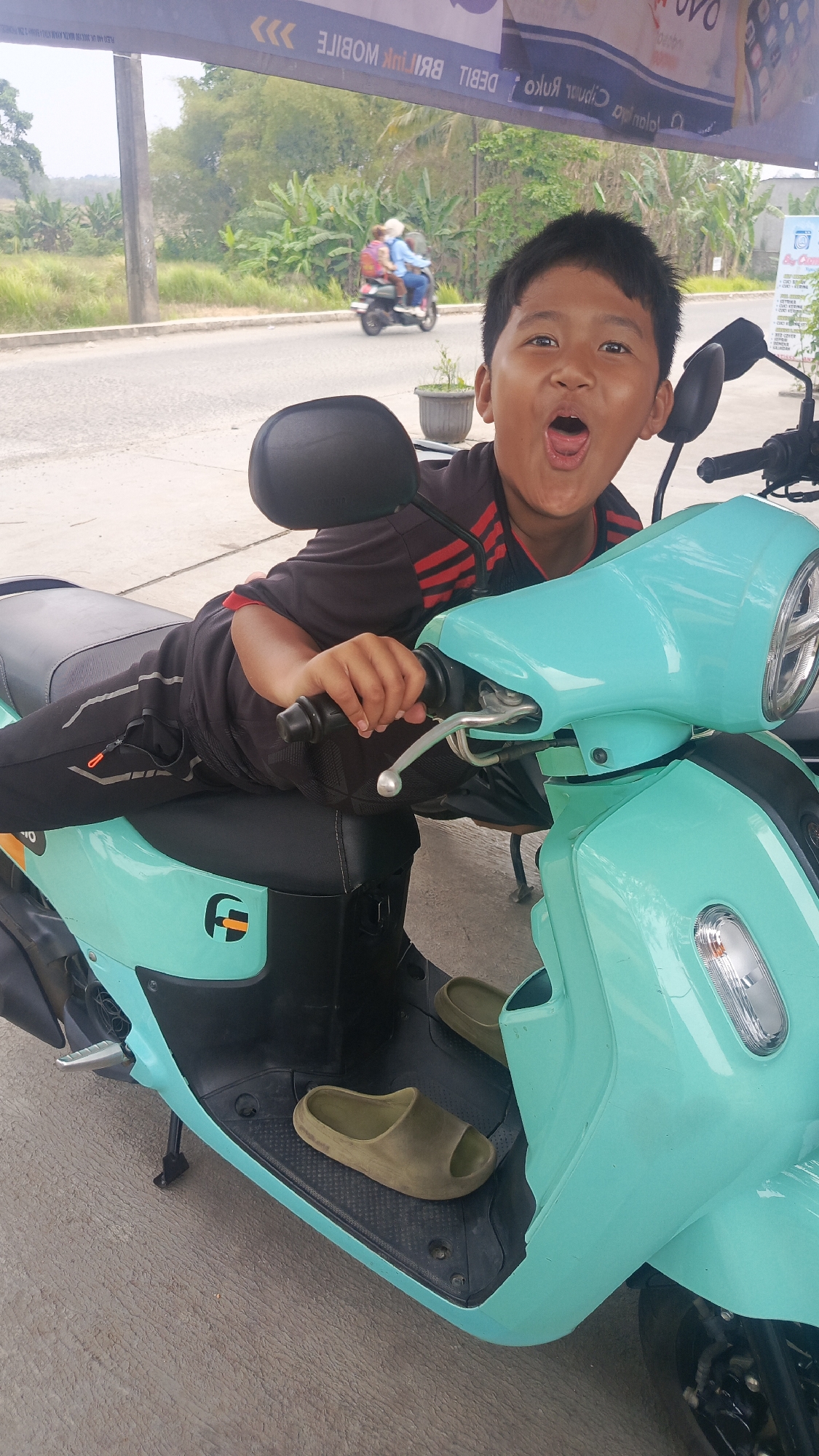 An Asian boy with black hair and an open mouth is lying on a turquoise electric scooter in Parung Panjang, Indonesia. He is wearing a dark t-shirt with red stripes on the sleeves and black pants. His right hand is on the scooter's handlebars. A pair of olive-green slides are placed on the scooter's floorboard. In the background, there is a road with a few trees and vegetation. A scooter with two people is visible on the road. A banner with text in an unknown language is hanging above.