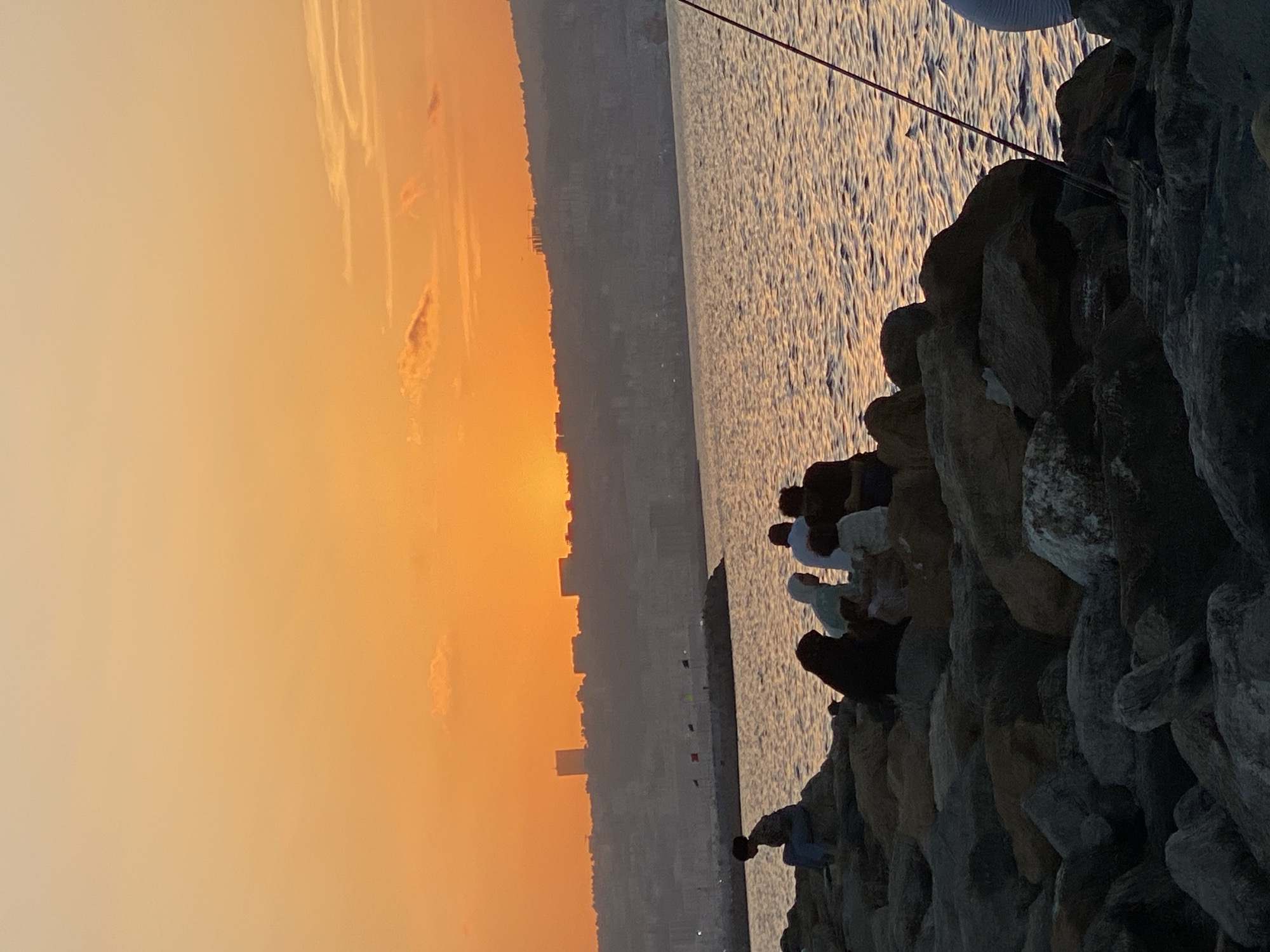 At sunset in Hussein Dey, Algeria, several individuals are gathered on a rocky pier. The sky is a vibrant orange, with the sun setting on the horizon, casting a warm glow over the scene. The city skyline is visible in the distance, silhouetted against the colorful sky. The water is calm, with gentle ripples reflecting the sunset. Some people are sitting on the rocks, while others are standing or walking. There is a fishing rod visible in the foreground, suggesting that some of the people may be fishing. The overall atmosphere is peaceful and serene, with a sense of community and relaxation.