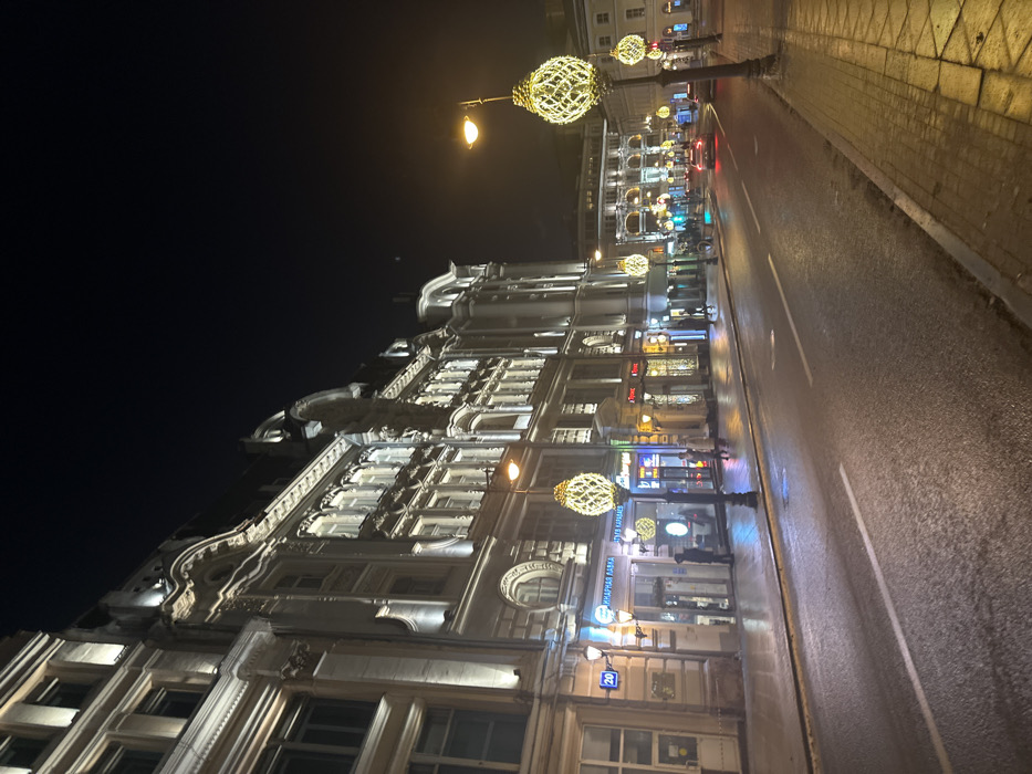 A nighttime street scene reveals a multi-storied building with ornate, lit windows and elaborate facade details. Decorative spherical streetlights illuminate the wet pavement and a narrow sidewalk. A road stretches alongside, reflecting lights from passing vehicles. Storefront signs are visible on the ground level of the building. The sky above is dark.