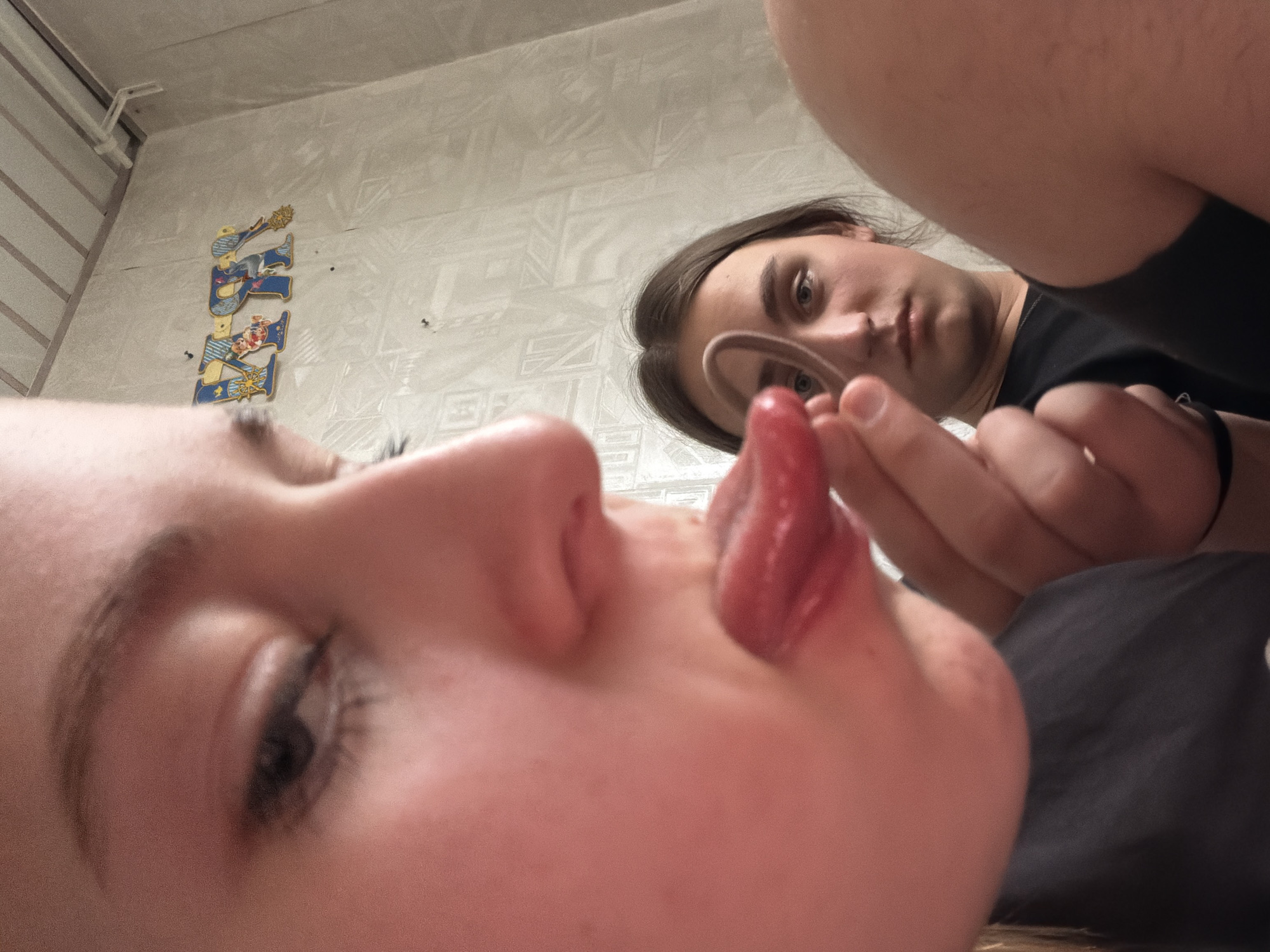 A close-up photograph depicts two individuals inside a room with patterned wallpaper and a tile-lined ceiling. One person, with fair skin and visible facial features including an eye, nose, and tongue, is the primary focus, situated very close to the camera. The tongue is extended. Another person is further away and out of focus. This person is holding a hair tie near the first person's mouth. There is a vertically oriented sticker on the wall behind the second person. The setting appears to be indoors, possibly a residential space in Perm, Russia, based on the general aesthetic.