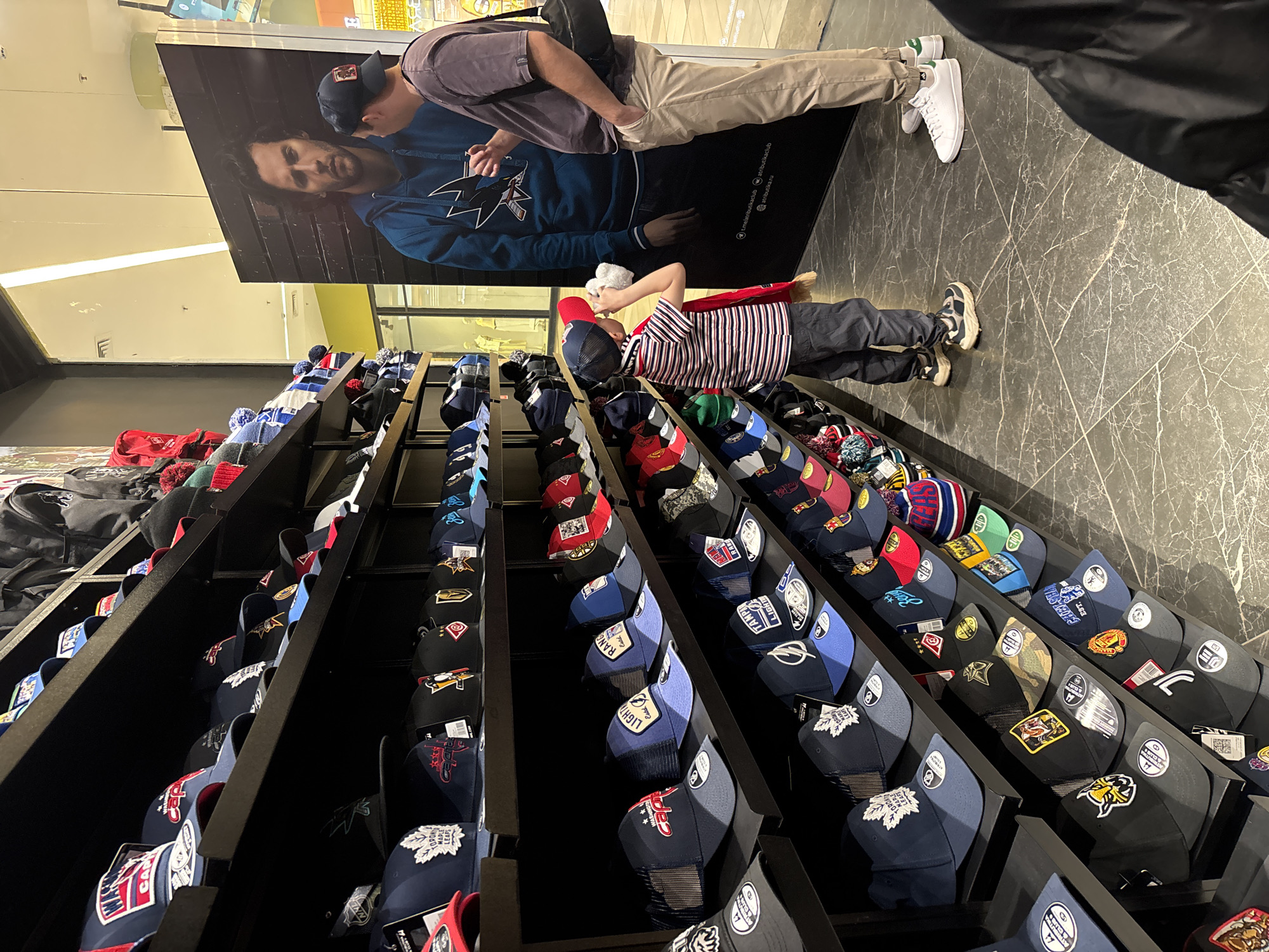 The image shows an indoor scene, likely inside a retail store or department store. The focus is on a display of baseball caps organized on shelves with a black frame. The caps feature various designs and logos.
A young boy, wearing a striped shirt and a red baseball cap, is looking at the display of hats. A man, dressed in beige pants, a grey shirt, and white sneakers, is standing next to a poster featuring a man in a blue sweatshirt. The man in beige pants seems to be examining something, possibly using his phone. The poster is placed behind the hat display.
The flooring appears to be made of dark marble or a similar patterned material. The lighting is artificial, typical of a retail environment.
Based on the OCR, there are URLs referencing "atributika.ru" and "tme/atributikaclub", which suggest that the scene might be located in Russia, possibly in the city of Rusanovo if that is indeed a relevant location in the context of the identified retail references.