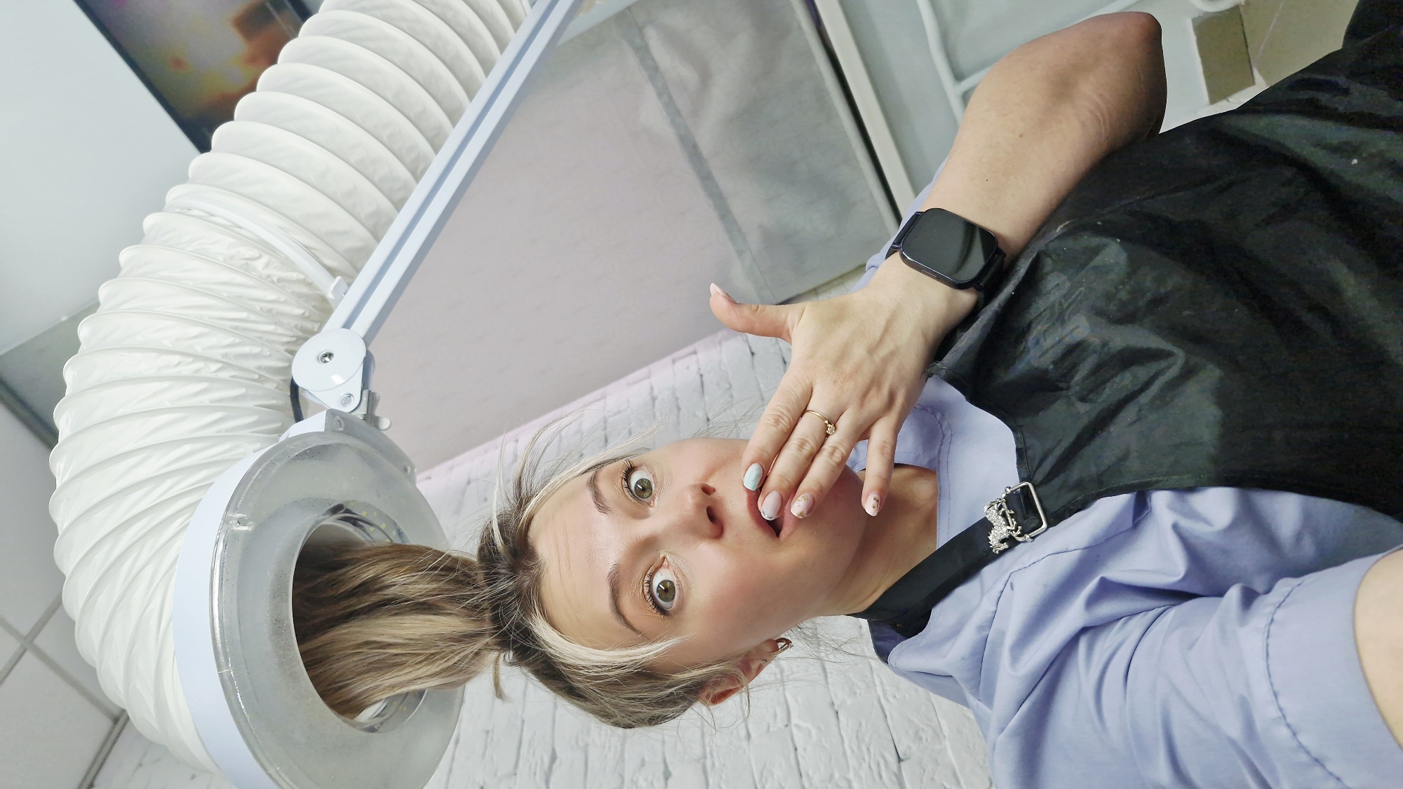 A close-up, low-angle shot captures a woman undergoing a hair treatment in what appears to be a salon in Otchor, Russia. Her light blonde hair, gathered into a bun, is inserted into the clear, circular head of a hair processing or drying device. This device features a prominent white flexible, corrugated tube extending upwards from its head.
The woman wears a light blue uniform-style top and a black protective cape or apron, secured by a metallic buckle with some embellishment. A black smartwatch is visible on her left wrist, and she wears a gold ring on the ring finger of her left hand. Her nails are manicured, featuring light blue and light pink polish with subtle decorative details.
She looks directly at the camera with wide, surprised eyes and a slightly open mouth, her left hand partially covering her mouth, conveying an expression of shock or playful disbelief. The background consists of a white brick wall, reinforcing the indoor salon setting. There is no visible text in the image.