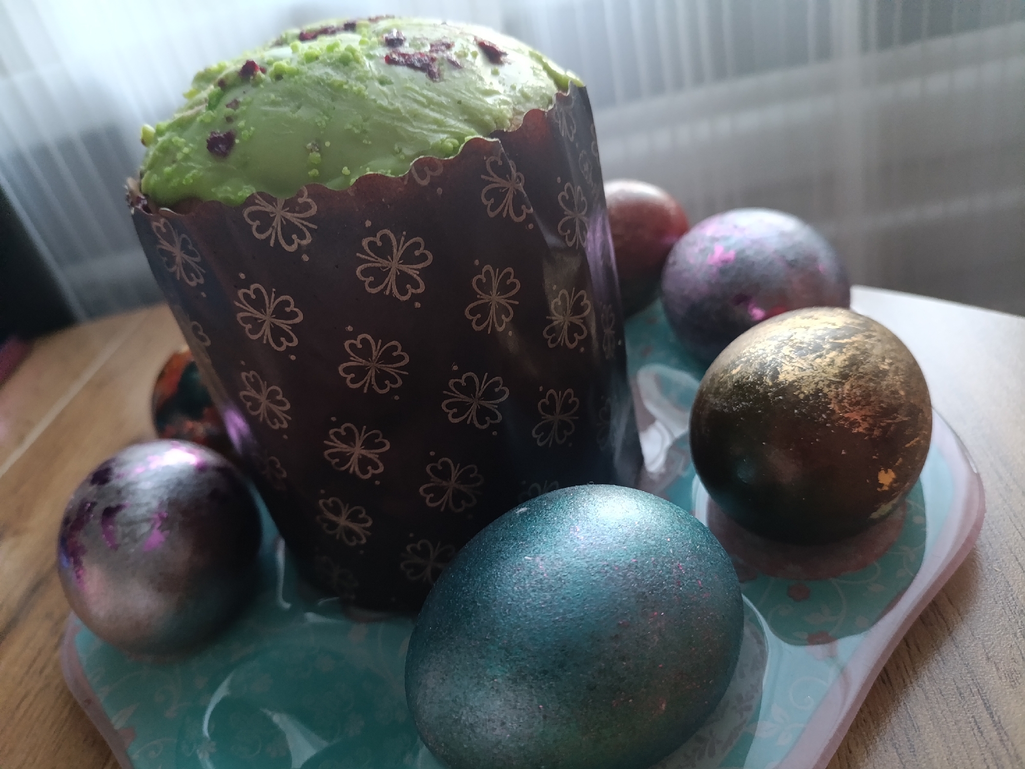 The image shows an Easter cake and painted Easter eggs on a blue and pink tray, resting on a wooden table. The Easter cake is in a dark brown paper baking form decorated with light brown clover patterns, and topped with green frosting. There are red sprinkles on the top of the frosting. Several Easter eggs surround the cake. The eggs are colored in metallic shades: gold, silver, teal, and bronze. There is a window with sheer white curtains in the background. The location and city of Izhevsk, Russia are not able to be definitively discerned from the visual information provided.