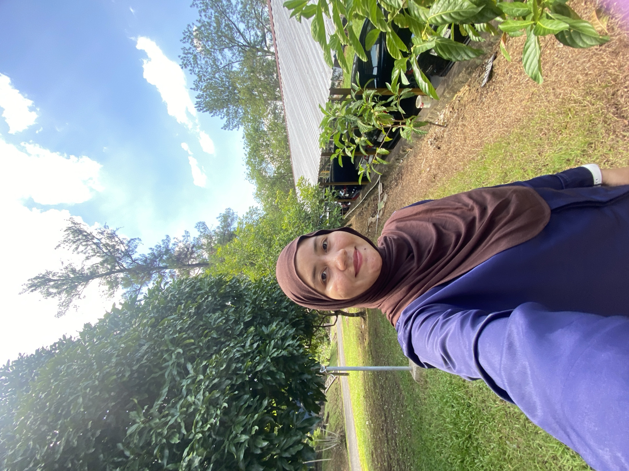 A woman wearing a brown hijab and a dark blue long-sleeved shirt takes a selfie outdoors. She is smiling and looking at the camera. The background shows a grassy area with trees and a building with a corrugated metal roof. Several cars are parked under a canopy. The location is identified as Banjarbaru, Indonesia. The sky is blue with scattered white clouds, and the sun is shining, casting shadows. The woman's skin shows some blemishes.