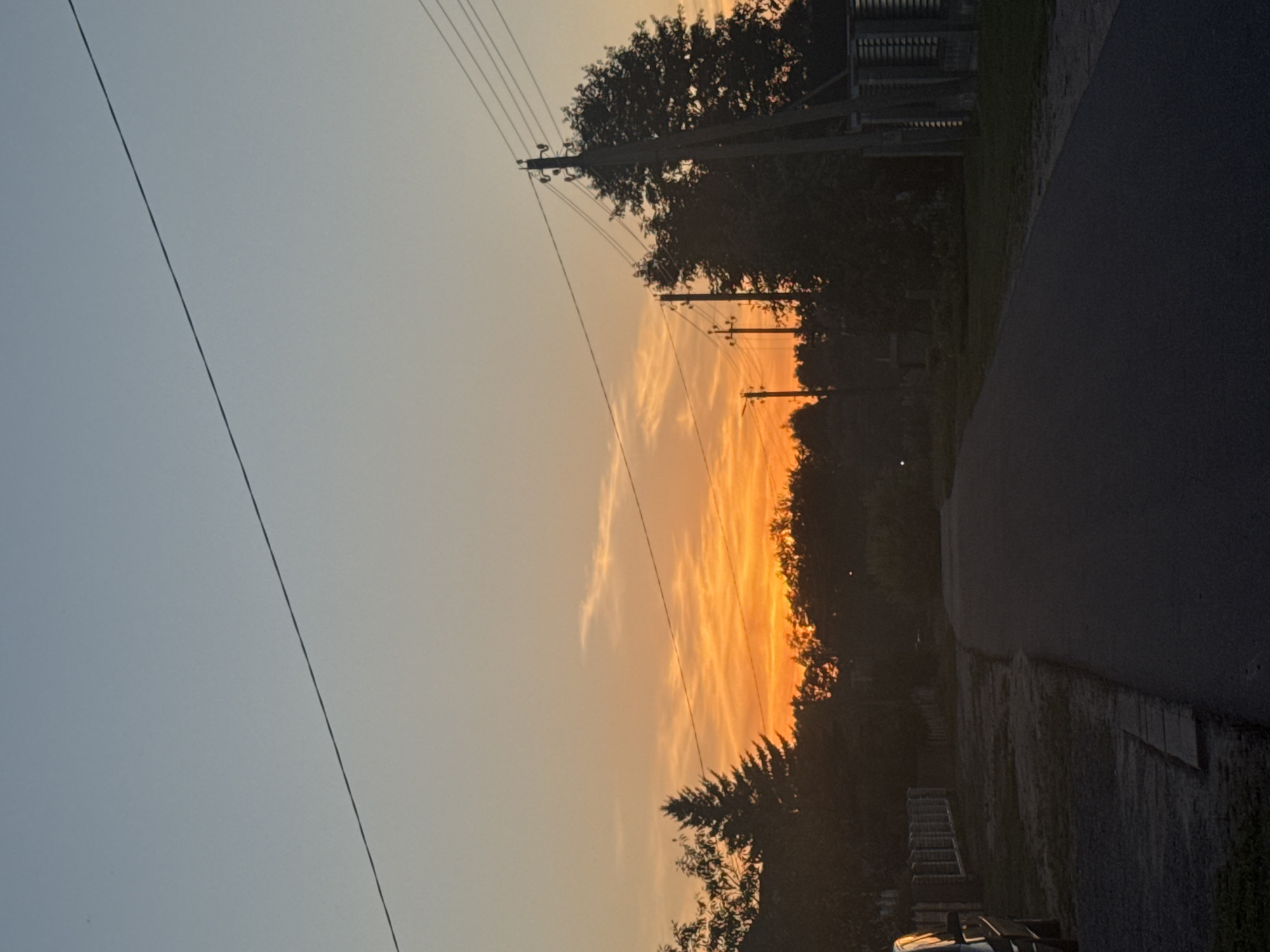 A photograph taken at dusk in Uzda, Belarus, depicts a rural road at the edge of a village. The sky is a gradient of soft blue at the top, transitioning to vibrant orange and yellow hues near the horizon, indicating a sunset. Wispy clouds are visible in the sunset area. Silhouetted against the sky are a line of trees, including a prominent conifer, and utility poles with overhead wires. The road, paved with asphalt, curves gently away from the viewer and into the distance. To the left of the road, a low fence made of brick pillars and horizontal wooden planks runs alongside a grassy area. A dark-colored vehicle is partially visible on the far left of the image. To the right of the road, more trees and a wooden utility pole are present, along with a glimpse of a structure that appears to be a building with corrugated metal siding. The overall scene conveys a sense of quiet and the end of a day in a countryside setting.