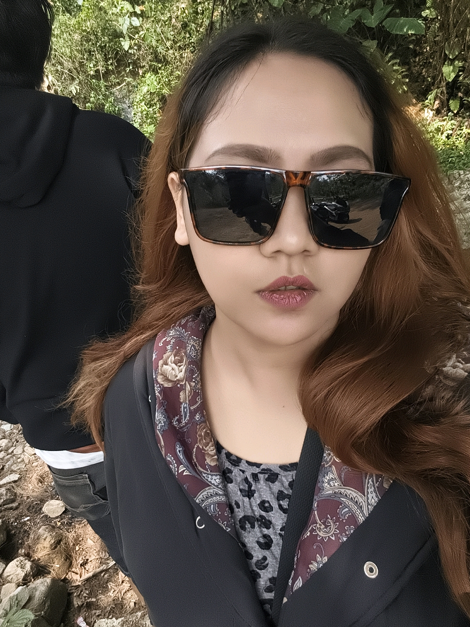 A young woman with auburn hair and sunglasses takes a selfie outdoors. She is wearing a black jacket over a leopard-print shirt with a paisley-patterned scarf. Her lips are painted a deep pink. Behind her, a person in a black hoodie is visible, and the background consists of lush green foliage and a rocky, outdoor path. The scene appears to be in a natural environment, possibly a park or a hiking trail in Babakan Madang, Indonesia.