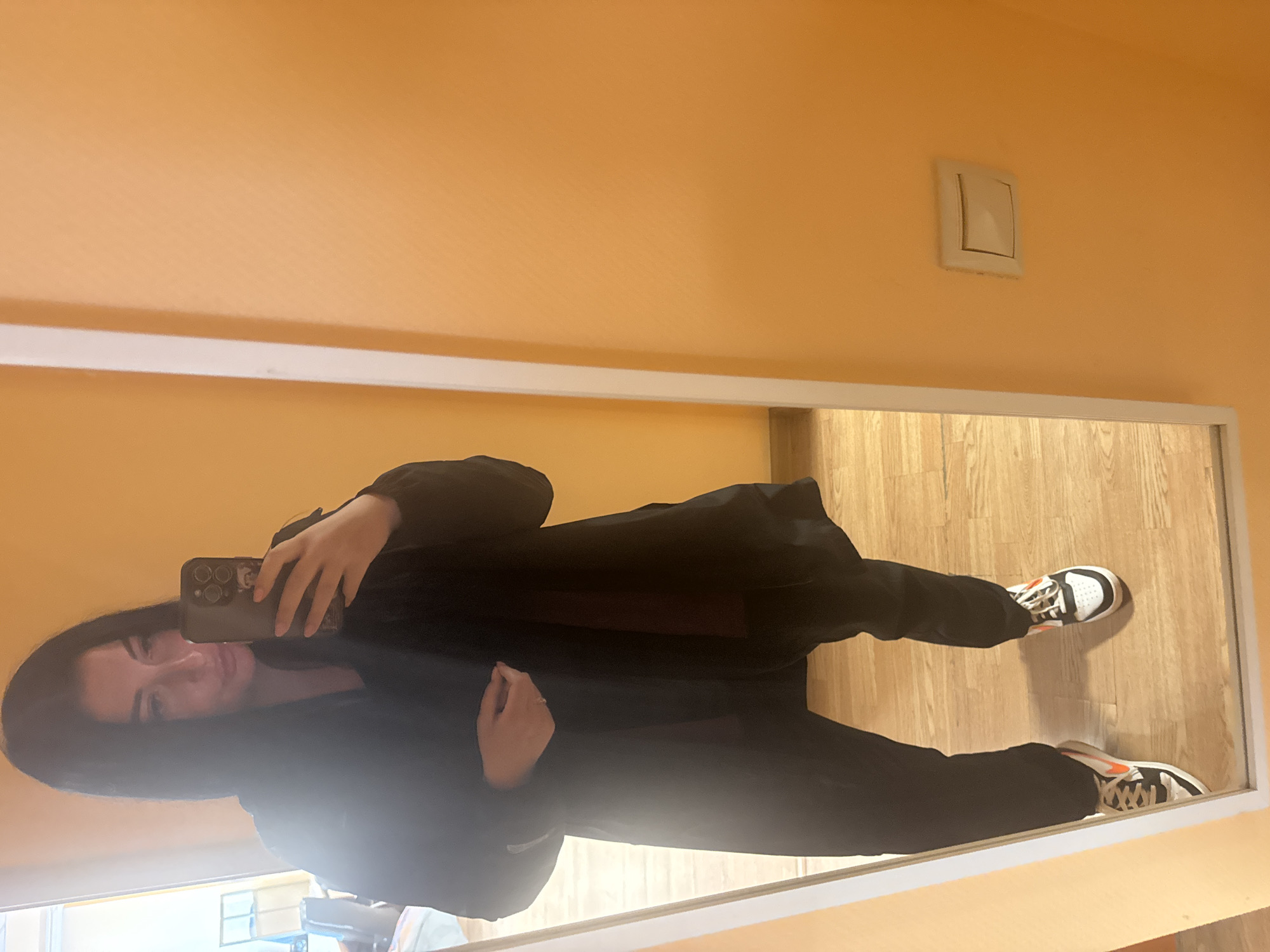 The image shows a full-body mirror selfie of a person taking a picture of themselves. The person is wearing a black jacket and black pants, along with white and black sneakers with orange accents. They are holding a smartphone in their right hand, with the camera pointed at the mirror to capture the reflection.
The setting appears to be indoors, with light orange walls and a wooden floor. A light switch is visible on the upper right wall. The lighting suggests it could be daytime, but it's difficult to determine the exact time or weather conditions. The person's facial expression is neutral, and the overall mood of the image is casual and everyday. The reflection shows the person from the waist down, including their legs and feet, and from the chest up, including their face and the phone they are holding. The image is oriented vertically, but the subject and the scene are presented sideways due to the camera angle.