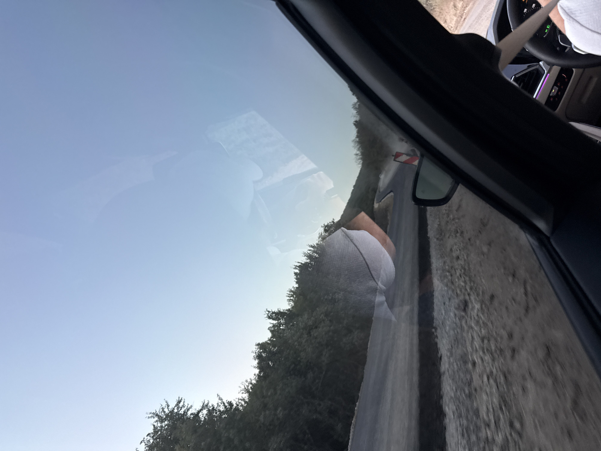 The image depicts a view from inside a car, looking out through the windshield. The scene appears to be a rural road under construction or repair, characterized by loose gravel and a partially paved surface. Trees line the left side of the road, forming a dense green backdrop. In the distance, the road curves sharply uphill, and there are signs of ongoing work, including a red and white striped barrier. The sky is a clear, pale blue, indicating it is likely daytime, possibly early morning or late afternoon given the soft light. Reflections are visible on the windshield, showing parts of the car's interior and possibly an object hanging or placed on it. A person's arm and hand are visible in the bottom right corner, suggesting the photographer is in the driver's seat. The overall impression is one of travel along a scenic, yet unfinished, route.