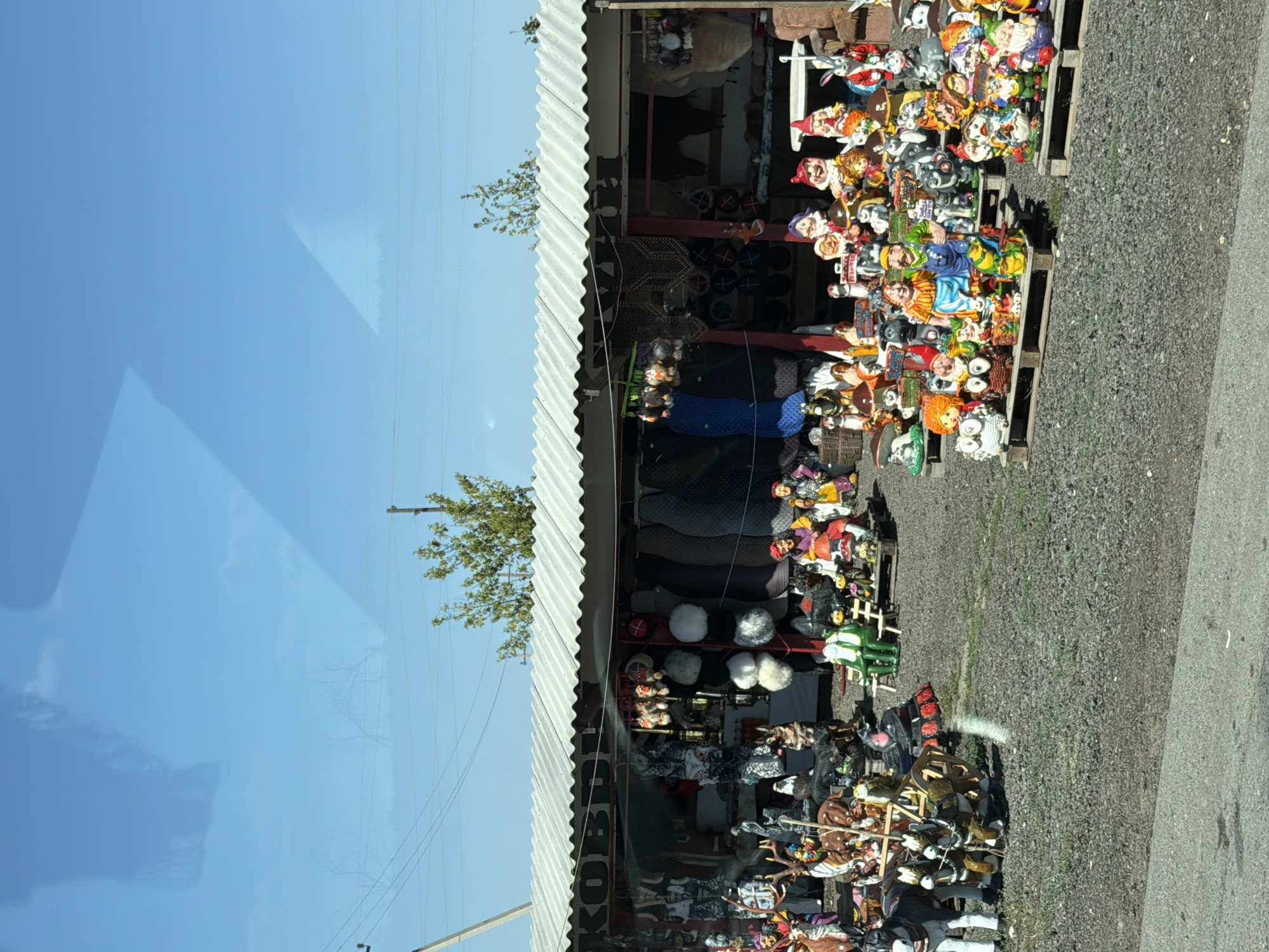 This image depicts an outdoor market stall in the city of Olenye, Russia, specializing in garden gnomes and other decorative figures. The stall is covered by a corrugated metal roof, supported by wooden posts. The merchandise is displayed on wooden pallets and the ground in front of the stall.
The stall is filled with numerous colorful garden gnomes, each with distinct facial features and attire, including pointed hats, beards, and varied clothing. Among the gnomes are other decorative figures such as rabbits, owls, deer, and a horse-drawn cart. Some figures are posed on mushroom-shaped bases, adding to the whimsical nature of the display.
Behind the figures, the stall's interior is visible, showcasing a variety of items including hats, coats, and other textiles hanging from the ceiling and walls. The ground around the stall is covered with gravel, and a paved road is visible in the foreground. The background features a clear blue sky with some reflection of clouds or objects from the viewer's perspective, and a few trees. The overall impression is that of a vibrant and diverse roadside attraction offering decorative garden ornaments.