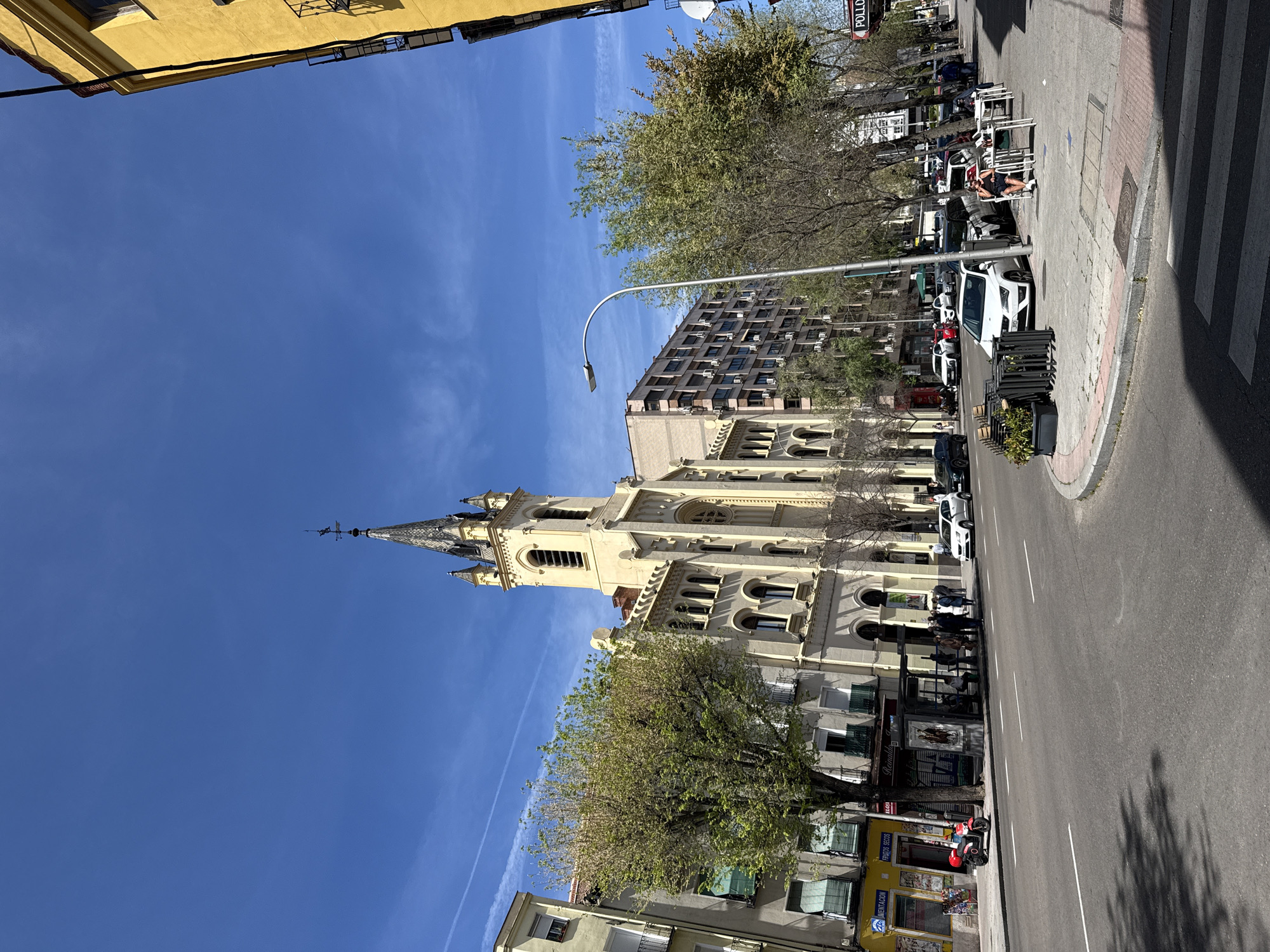 The image shows a street scene in Madrid, Spain. The most prominent feature is a tall, light-colored church with a pointed steeple. Buildings line both sides of the street, with trees interspersed. Cars are parked along the curb, and people are walking on the sidewalk and sitting at outdoor cafes. The sky is clear and blue.