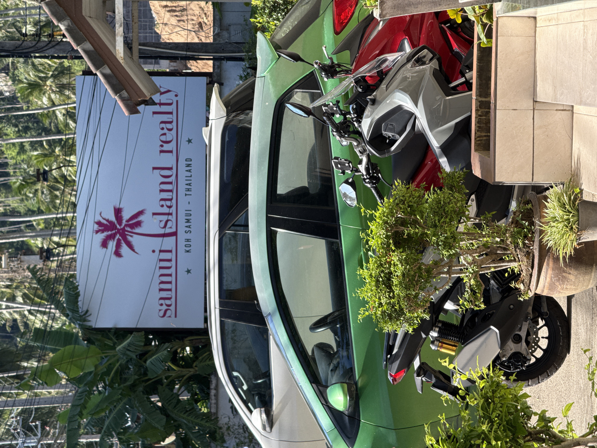An outdoor, daytime scene in Koh Samui, Thailand, features a white SUV parked behind a bright green car. A pink palm tree logo is displayed on a white sign above the vehicles, with the text "samui island realty" and "KOH SAMUI - THAILAND" visible. In the foreground, two motorcycles are parked: one is gray and the other is red, partially obscured by greenery in terracotta pots. The background consists of lush green trees and foliage.