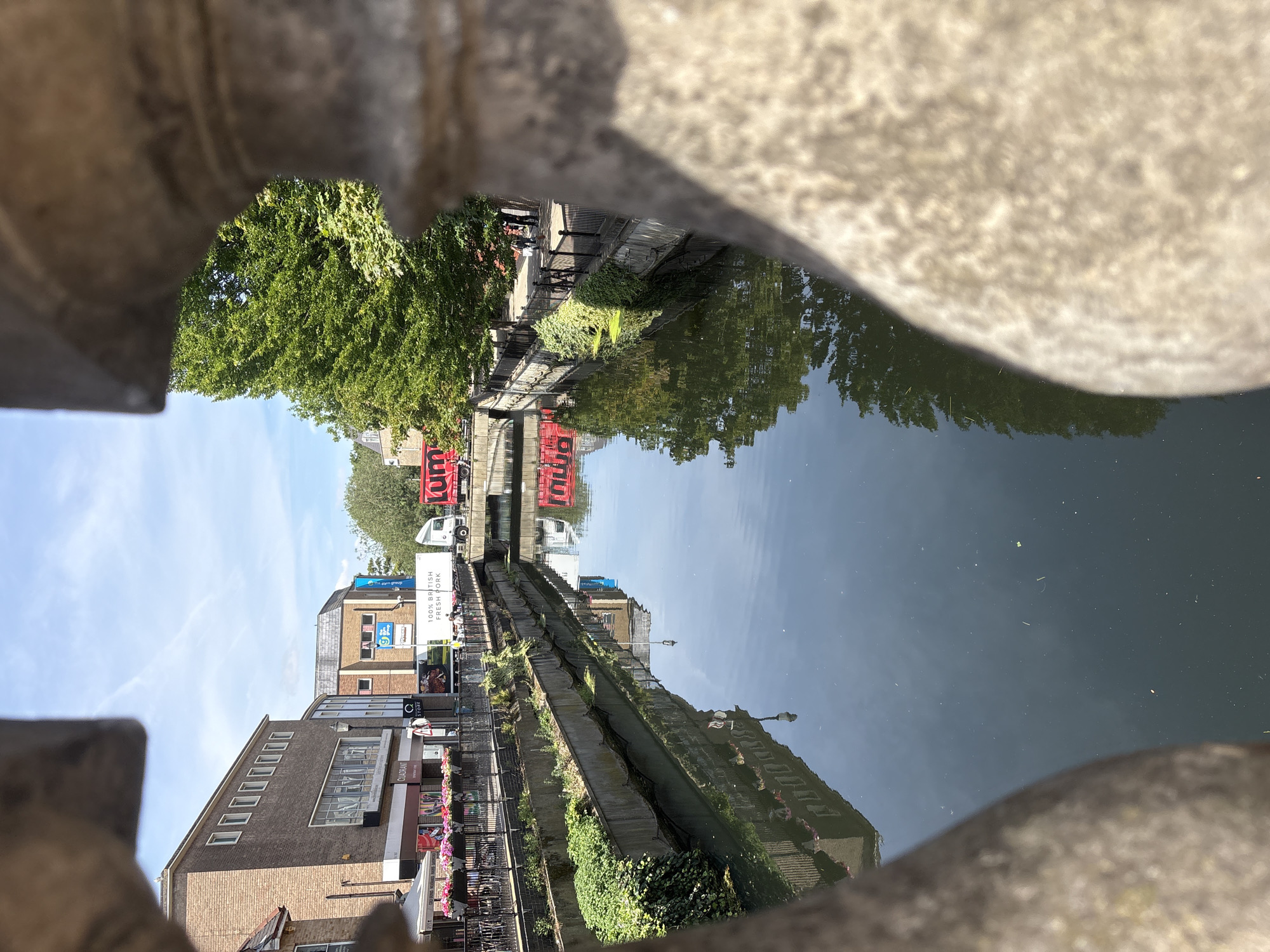 This is a photograph taken from a bridge overlooking a canal or river in an urban environment. The foreground is dominated by the blurred texture of a stone balustrade, suggesting a viewpoint from within a decorative architectural element of the bridge. The canal water is dark and reflective, mirroring the buildings and trees on either side.
On the left bank, a brick building with multiple windows and a glass-fronted ground floor housing shops can be seen. Above the shops, there is signage including "QUADRANT" and what appears to be a blue advertisement for "Go Group". Planters filled with pink and red flowers line the edge of the walkway above the canal.
On the right bank, a lush green tree is prominent, its branches reaching towards the sky. Beyond the tree, more buildings are visible, and a black metal railing runs along the edge of the walkway, separating it from the canal. Several people are walking along this right-hand path.
In the middle of the canal, two red trucks are visible. The one closer to the foreground has the word "NUM" printed on its side in large white letters. Further down the canal, another, possibly identical, red truck is partially visible. The sky above is blue with scattered white clouds. The overall scene depicts a sunny day in a town or city with a prominent waterway.