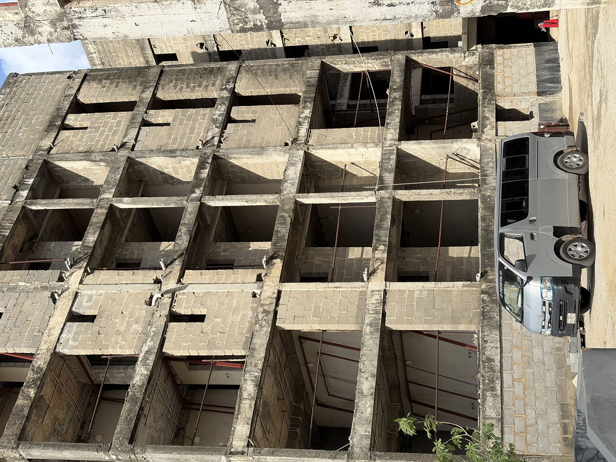An outdoor shot depicts a derelict, multi-story building under construction, partially revealing the sky. The building's façade is characterized by evenly spaced, rectangular, unfinished window openings, creating a grid-like pattern. Various pipes and fixtures are visible in the raw interiors of the building. A gray Toyota HiAce van is parked adjacent to the building. The environment seems urban, and the location is within the city of Lagos, Nigeria.