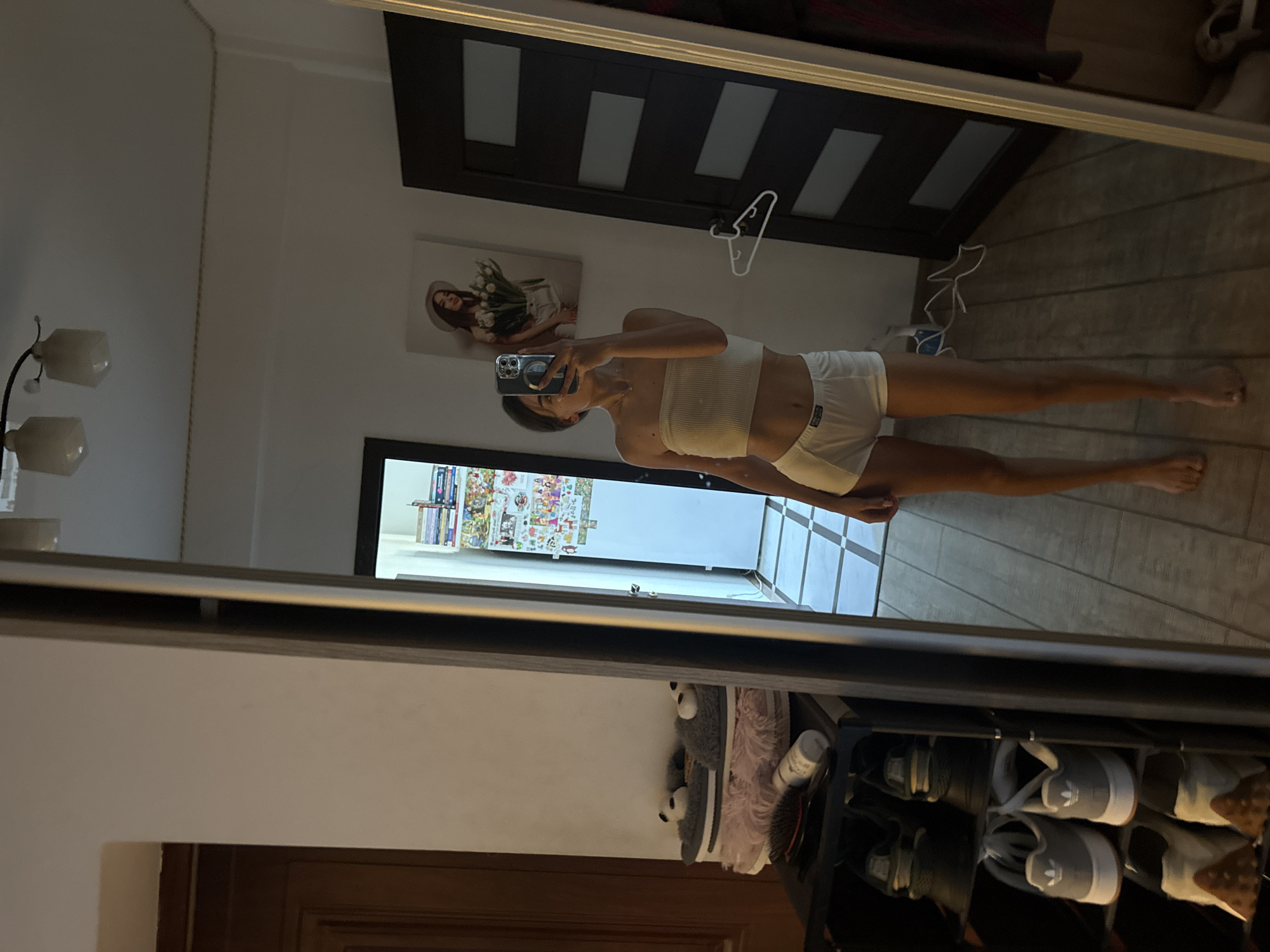 A young woman is taking a mirror selfie in a room. She is wearing a white tube top and white shorts. She is standing in front of a large mirror, which reflects the room. To her left, there is a shoe rack with several pairs of shoes. To her right, there is a dark door with rectangular panels. Above the door, there is a white hanger. On the wall behind her, there is a framed picture of a woman wearing a hat and holding flowers. The room appears to be a bedroom or dressing area, with tiled flooring and wooden doors. The overall impression is of a personal, domestic space, likely within a residential building. The presence of a shoe rack and what appears to be a closet further suggests a living space. There are no immediate indicators of the specific location within Chișinău, Moldova, as the image lacks any distinctive landmarks or signage.