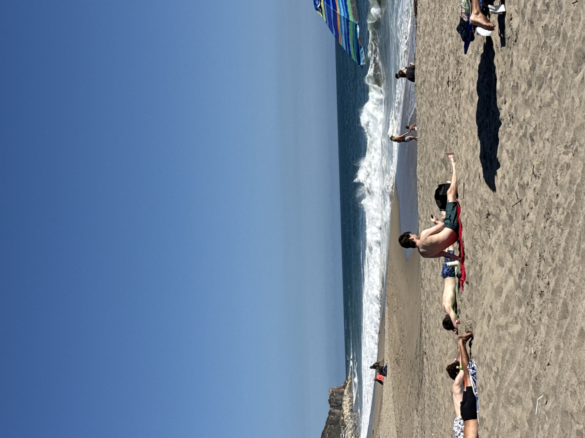 The image depicts a sunny day at a beach in Malibu, California, USA. The sky is a clear, vibrant blue, with a distant coastline featuring rocky cliffs on the left. The ocean is active with moderate waves breaking on the shore. In the foreground, several individuals are sunbathing on the sandy beach. One person is lying on their stomach on a red towel, while another is lying on their side. Further back, two individuals are seated. A striped beach umbrella is visible on the right side of the frame. The overall scene conveys a relaxed atmosphere of a typical beach day.