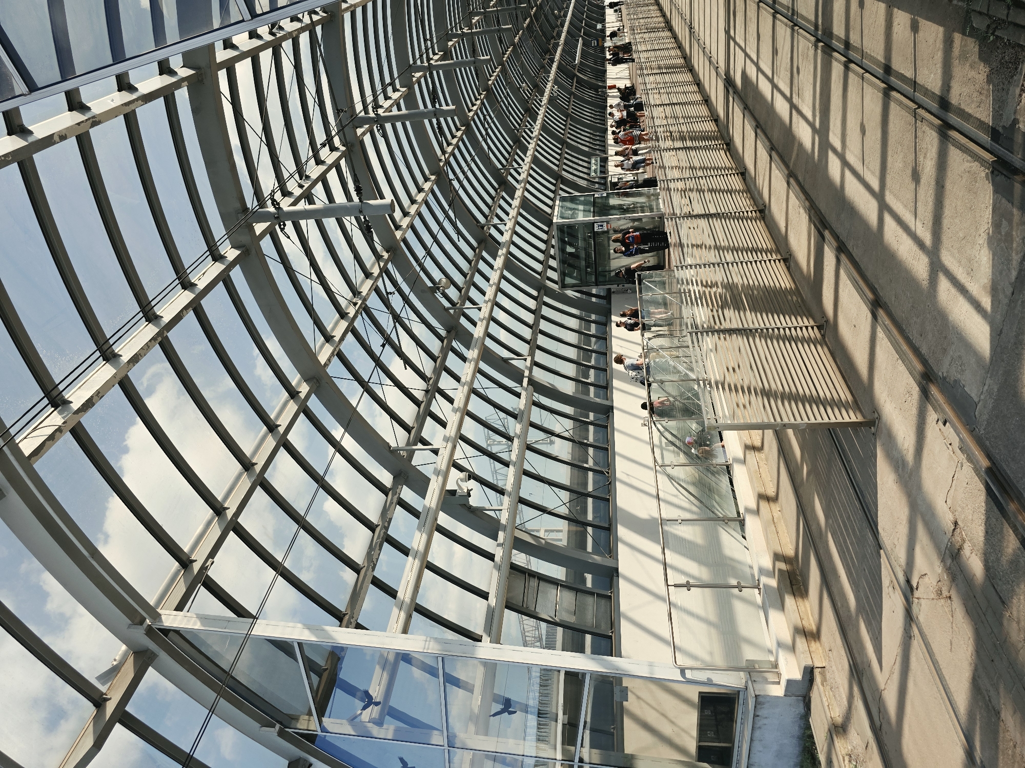 This image depicts the Poznan Glowny train station's modern glass and steel roof structure, which spans over the platforms and waiting areas. The arched roof is supported by a series of grey metal beams and is made of translucent glass panels, allowing natural light to filter through. The sky, with scattered white clouds, is visible through the roof.
On the left side of the image, a glass-enclosed elevator shaft is visible. In the background, people are seen walking and standing on the platform. A silver railing with glass panels separates the platform from the tracks. The ground in front of the platform is paved with concrete, and train tracks run along the lower part of the image. Sunlight casts long, geometric shadows from the roof structure onto the platform and tracks.
Several individuals are visible as silhouettes or partially obscured figures, engaged in activities such as waiting, walking, and ascending an escalator. The overall impression is one of a bustling, contemporary transportation hub.
