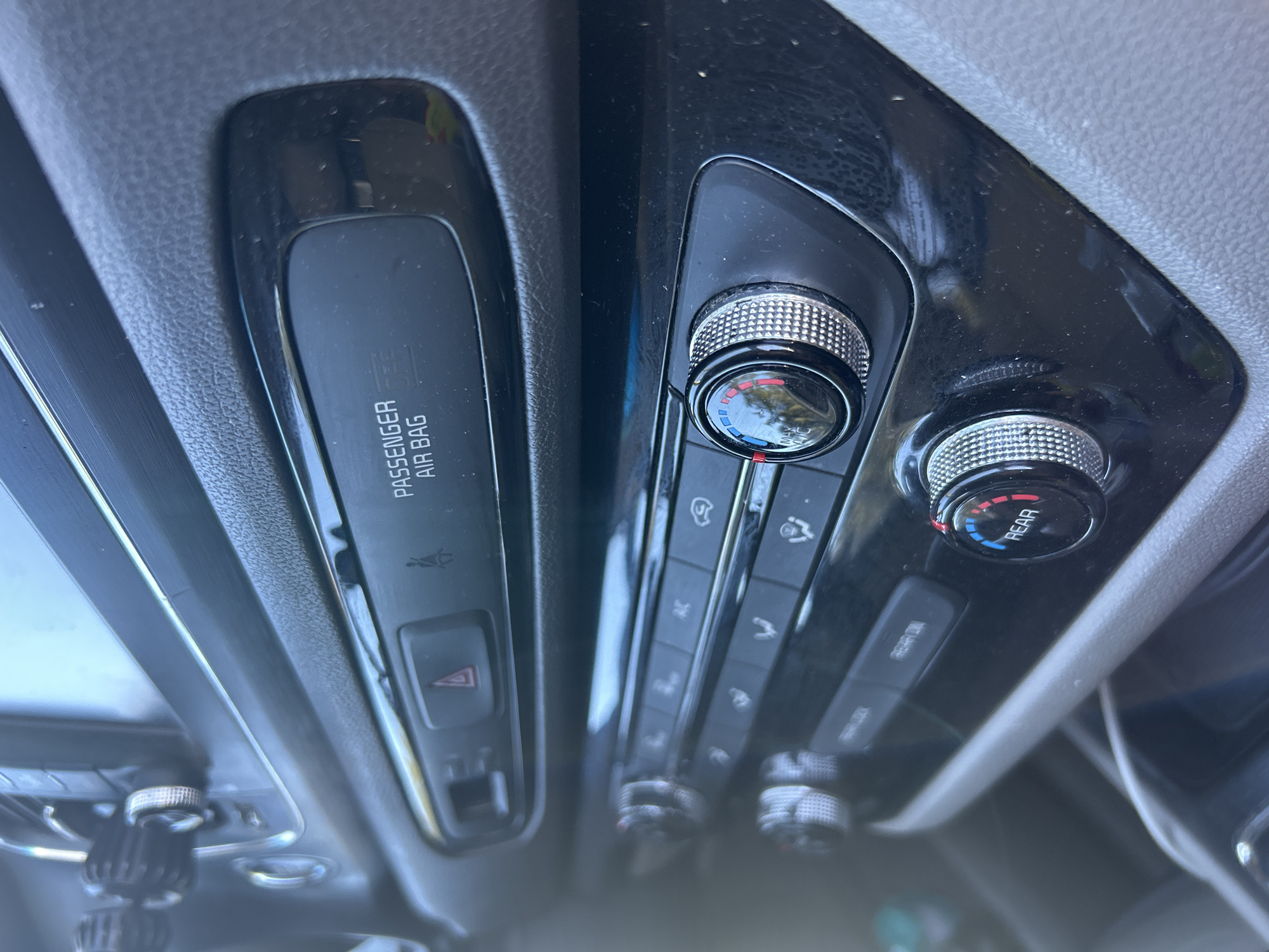 The image shows a close-up of a car's climate control panel. There are two rotating dials with segmented rings of red and blue to indicate temperature settings. Above the dials, there are buttons with various icons and text labels, including "PASSENGER AIR BAG" and a hazard light symbol. The panel is made of black plastic with a glossy finish, and some dust is visible on the surface. The car appears to be parked, with a blurred background that suggests an outdoor environment, possibly a parking lot or street. The lighting suggests it is daytime. The overall impression is that of an interior car shot focusing on the dashboard controls. The location in the city of Everett, United States of America, is not directly discernible from the visual information in the image.