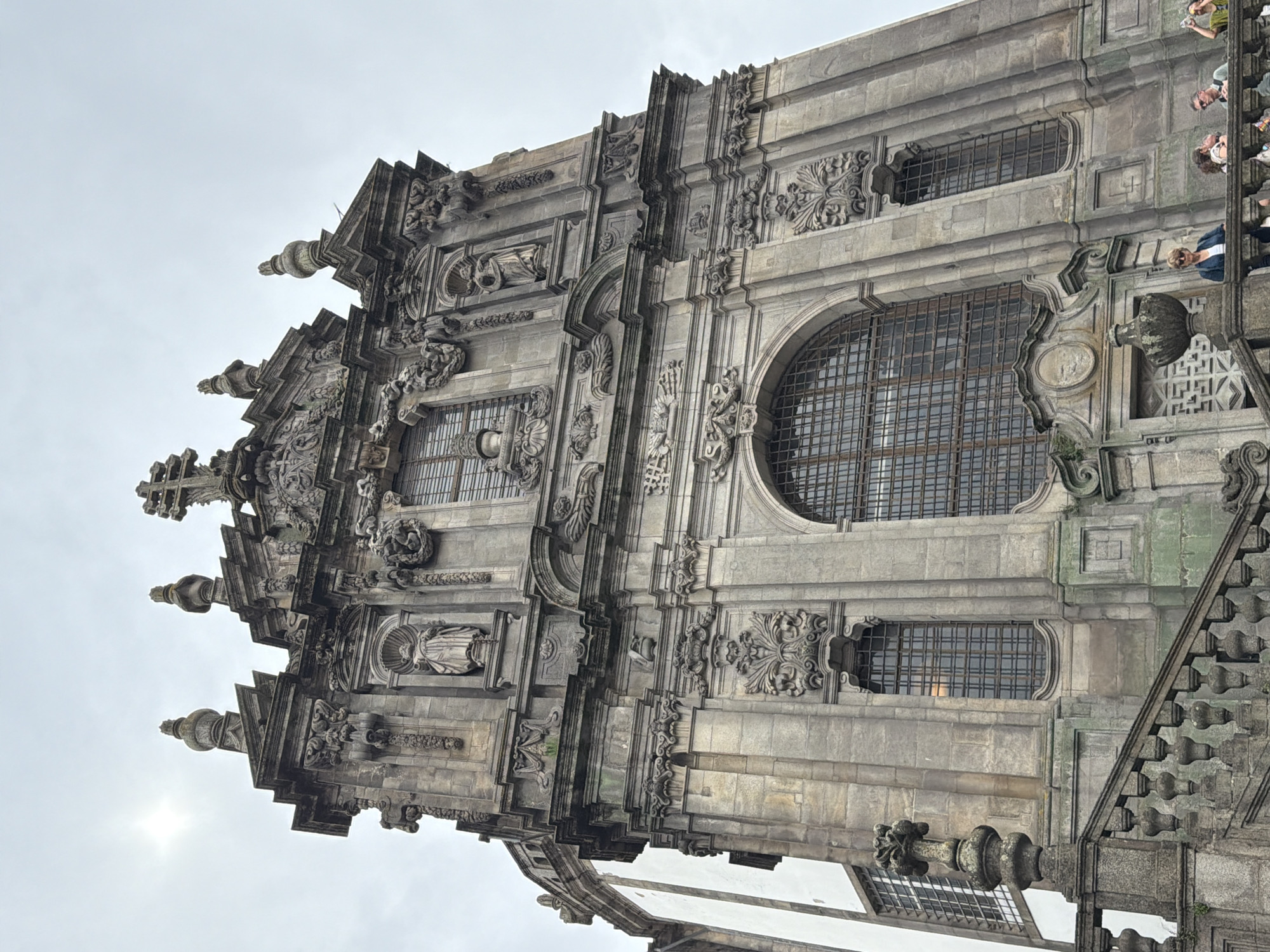 The image shows the exterior of a grand, ornate Baroque building, identified as the Clérigos Church in Porto, Portugal. The building is constructed of grey stone and features intricate architectural details, including statues, carvings, and large arched windows with metal grates. The perspective is looking upwards from a stone staircase that leads towards the entrance of the building. The sky is overcast. In the lower right portion of the image, several people are visible standing on the staircase and an adjacent railing, observing the building. They are casually dressed, and their interactions appear to be social.