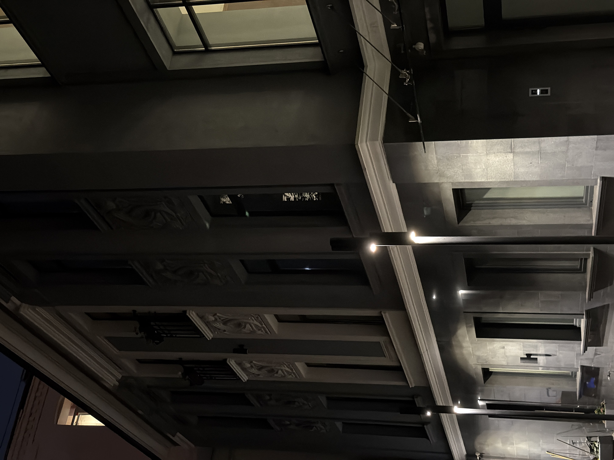 The image shows a nighttime view of a modern building exterior with architectural details, possibly in Moscow, Russia. The building facade is dark gray with lighter gray stone cladding on the lower levels. The upper floors feature decorative relief sculptures above windows. Several tall, thin streetlights illuminate the lower portion of the building. The windows are dark, with some reflecting light from within or outside. A small, light-colored intercom or call button is visible on a dark section of the building to the right. The scene suggests an urban environment at night, with the building appearing to be an office or residential complex.