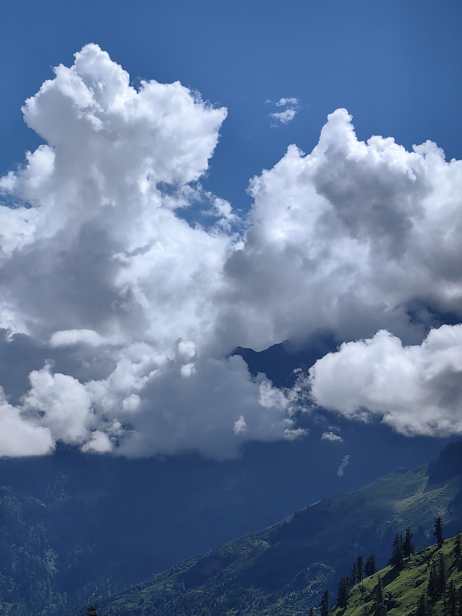 The image captures a dramatic sky over mountainous terrain in Kulang, India. Dominated by a vivid blue sky, the scene is filled with voluminous, textured white clouds, some casting shadows that create depth. The mountains below are shrouded in a dark, atmospheric haze, with visible slopes of lush green vegetation and scattered pine trees, particularly on the right side of the frame. The upper portion of the image focuses on the clouds, while the lower portion reveals the detailed topography of the mountains.