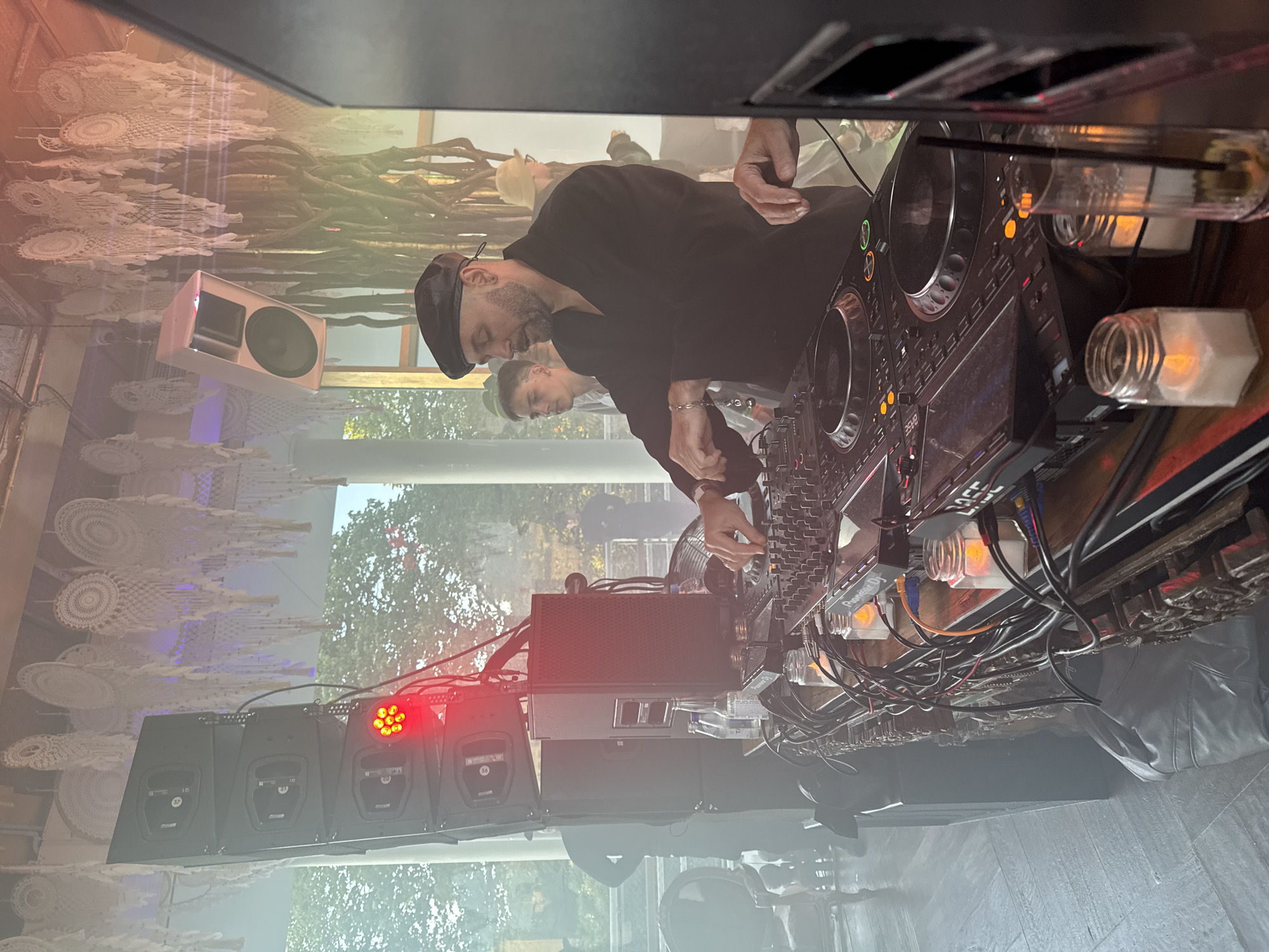 The image depicts a DJ performing at an outdoor event, possibly a festival or party. The DJ, a man wearing a black beret and a black shirt, is actively mixing on a Pioneer DJ setup. Several speakers are visible, including a large stack of black speakers on the left and a white speaker hanging from above on the right. The background shows trees and possibly a building with an open-air structure. The scene is illuminated by ambient lighting, with some smoke or haze present, adding to the atmosphere. In the foreground, there are candles in jars and a drink. Other people are blurred in the background, suggesting a lively gathering. The overall setting appears to be informal and celebratory.