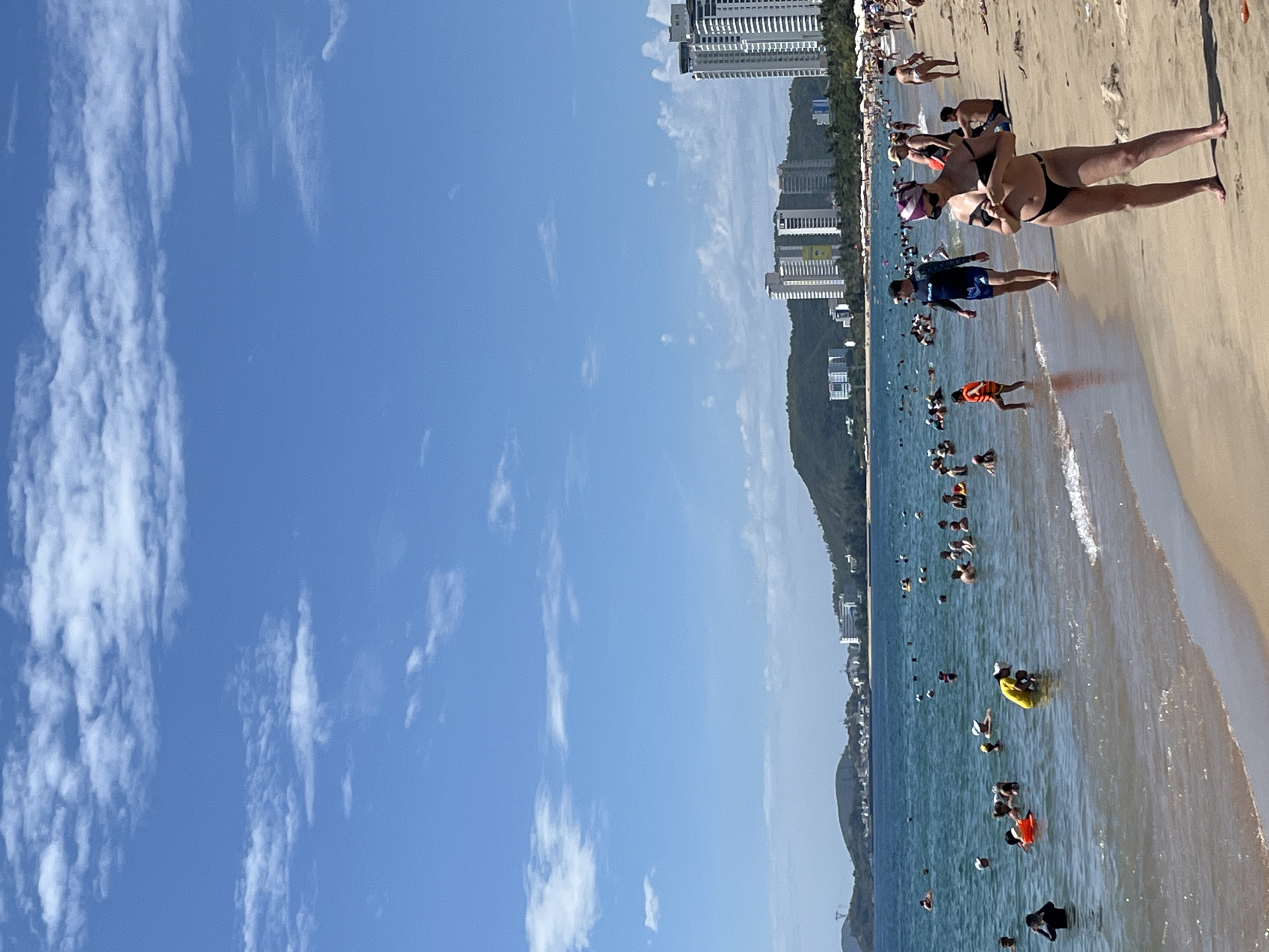 The image depicts a crowded beach scene in Nha Trang, Vietnam. In the foreground, a woman in a black bikini stands on the sand, facing left, with her hands adjusting her bikini top. Behind her, a man in blue swim trunks and a dark rash guard walks towards the water. Further out, numerous people are enjoying the ocean, with some swimming and others wading. An orange life vest is visible on a young girl in the water. The sea is a clear blue, with gentle waves lapping the shore. In the background, a line of tall, modern buildings stands against a backdrop of lush green hills. The sky is a vibrant blue, dotted with white, fluffy clouds. Beach umbrellas and lounge chairs are scattered along the sandy shoreline, indicating a popular tourist destination.