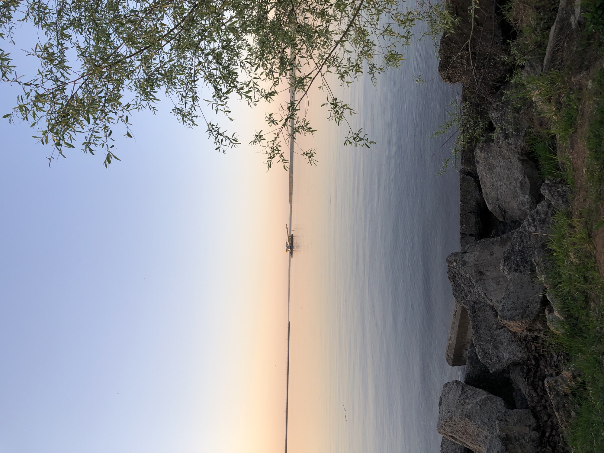 The image depicts a body of water, likely the Kama River in Chaikovsky, Russia, at sunset. The sky transitions from blue to a warm orange near the horizon. A dredging barge is visible on the water. In the foreground, there are rocks and vegetation on the right side of the image, and tree branches with leaves in the upper left corner. The water is calm with subtle ripples. The orientation of the image is rotated 90 degrees clockwise.