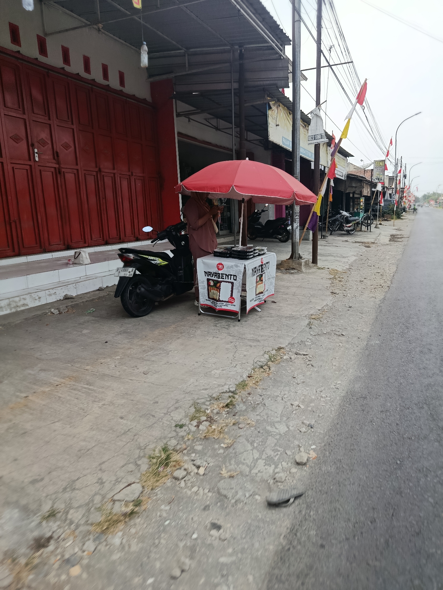 The image captures a street scene in Bantarsari, Indonesia, with a focus on a small vendor stall. In the foreground, a paved sidewalk runs alongside a road. A black motorcycle is parked on the sidewalk. Next to the motorcycle is a small, white table with a red umbrella overhead. On the table are several black boxes, likely food containers, with a "NAVABENTO" logo visible. A person, partially obscured by the umbrella, appears to be standing behind the table, possibly the vendor. To the left of the stall is a building with a red, corrugated metal facade and a white wall above it. The street itself is paved, and further down the road, other buildings and more motorcycles are visible. Several Indonesian flags are displayed along the street, suggesting a festive occasion or national holiday. The sky is overcast.