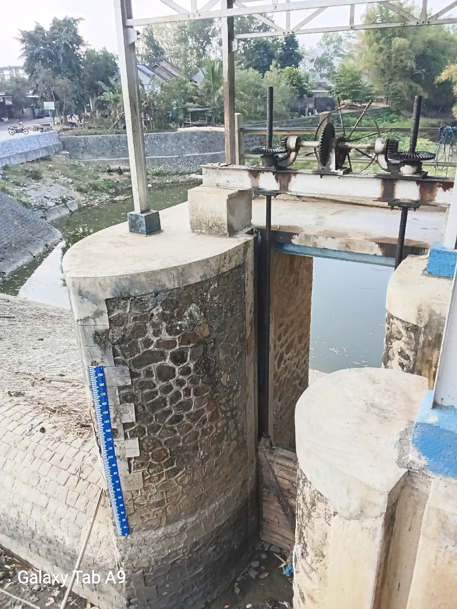 The image depicts a water sluice gate system at a canal in Sumberingin, Indonesia. The foreground features a large, curved concrete structure with a stone facade, supporting the sluice gate mechanism. A vertical blue and white water level gauge is affixed to the stone wall, showing markings from 0 to 190. Beyond the sluice gate, a body of water is visible, with a wooden gate mechanism visible in the background on the right side of the image. The surrounding environment includes a canal with water on the left, lined with concrete and stone embankments. In the distance, lush green trees and vegetation are present, along with some buildings and what appears to be a road with a motorcycle. The overall scene suggests an operational water management facility.