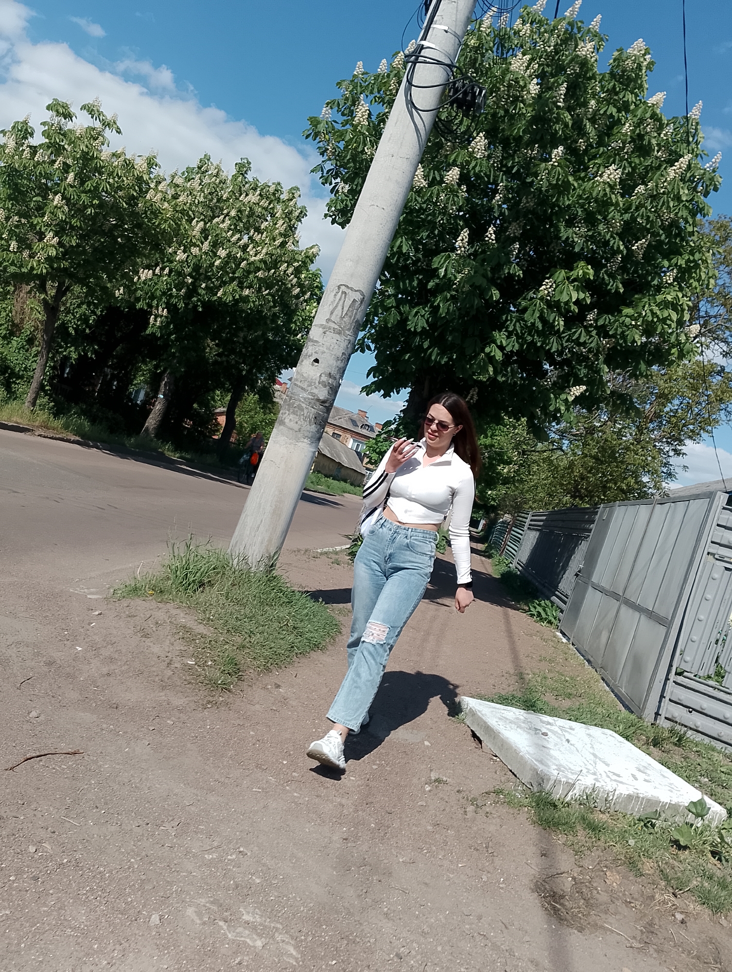 A full-shot photograph captures a young woman walking outdoors on a sunny day in Unknown, Unknown. She is wearing a long-sleeved white crop top, ripped blue jeans, and white sneakers. Sunglasses are perched on her face, and she carries a white bag over her shoulder.
The woman is walking on a dirt path adjacent to a paved road. A leaning concrete utility pole stands to her left, with electrical wires extending from its top. Lush green trees with white blossoms surround the scene, and a gray metal fence runs along the right side of the path. A large, flat concrete slab rests on the ground near the fence. The sky above is blue with scattered white clouds. The scene is tilted slightly to the right.