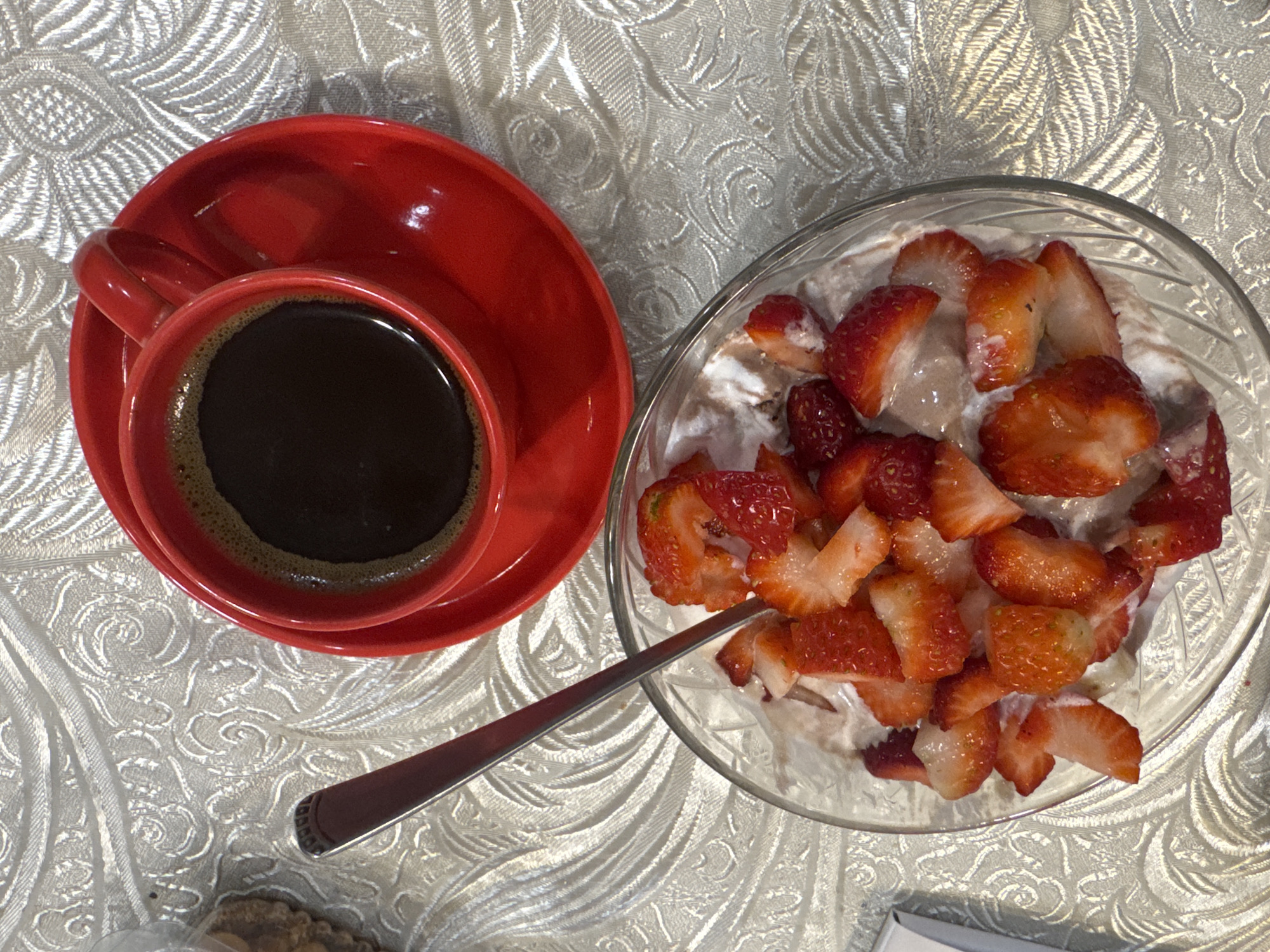 The image shows a red cup of coffee on a saucer, next to a glass bowl filled with what appears to be sliced strawberries mixed with yogurt or cream. A silver spoon rests on the patterned tablecloth between the cup and the bowl. The scene is indoors, likely on a table. The location is Yerevan, Armenia.