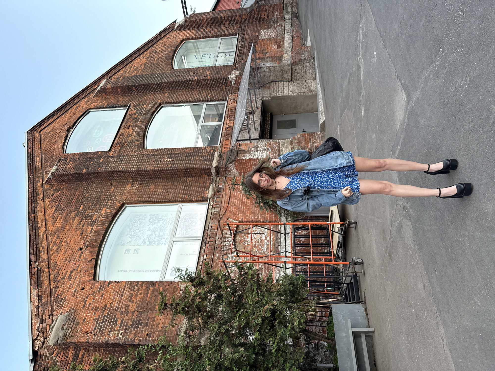 The photograph shows a woman standing in front of a brick building. The woman is wearing a blue floral dress and a denim jacket, and she has long brown hair and glasses. The building has arched windows, some with text or graphics visible inside. The photograph appears to be taken at the Wine Factory in Moscow, Russia, based on the architectural style and details of the brick building. There is a tree and some metal structures in the foreground, and a paved surface.