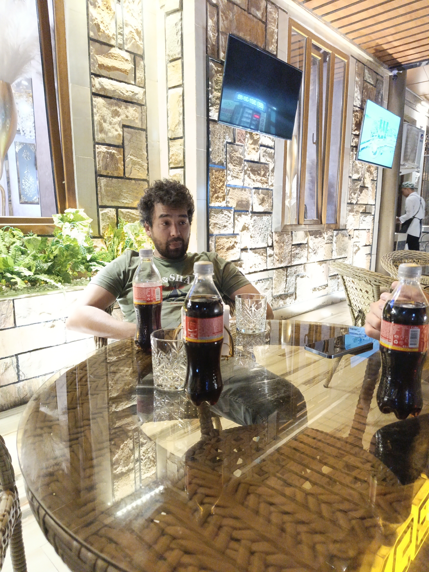 A man with curly hair is sitting at a round glass table. He is wearing a green t-shirt. On the table are two bottles of Coca-Cola and a glass of water. A phone is also on the table. In the background, there are stone walls, plants, and a television screen displaying what appears to be a clock. There is another person in the background wearing a white uniform. The scene appears to be indoors, possibly a restaurant or cafe.