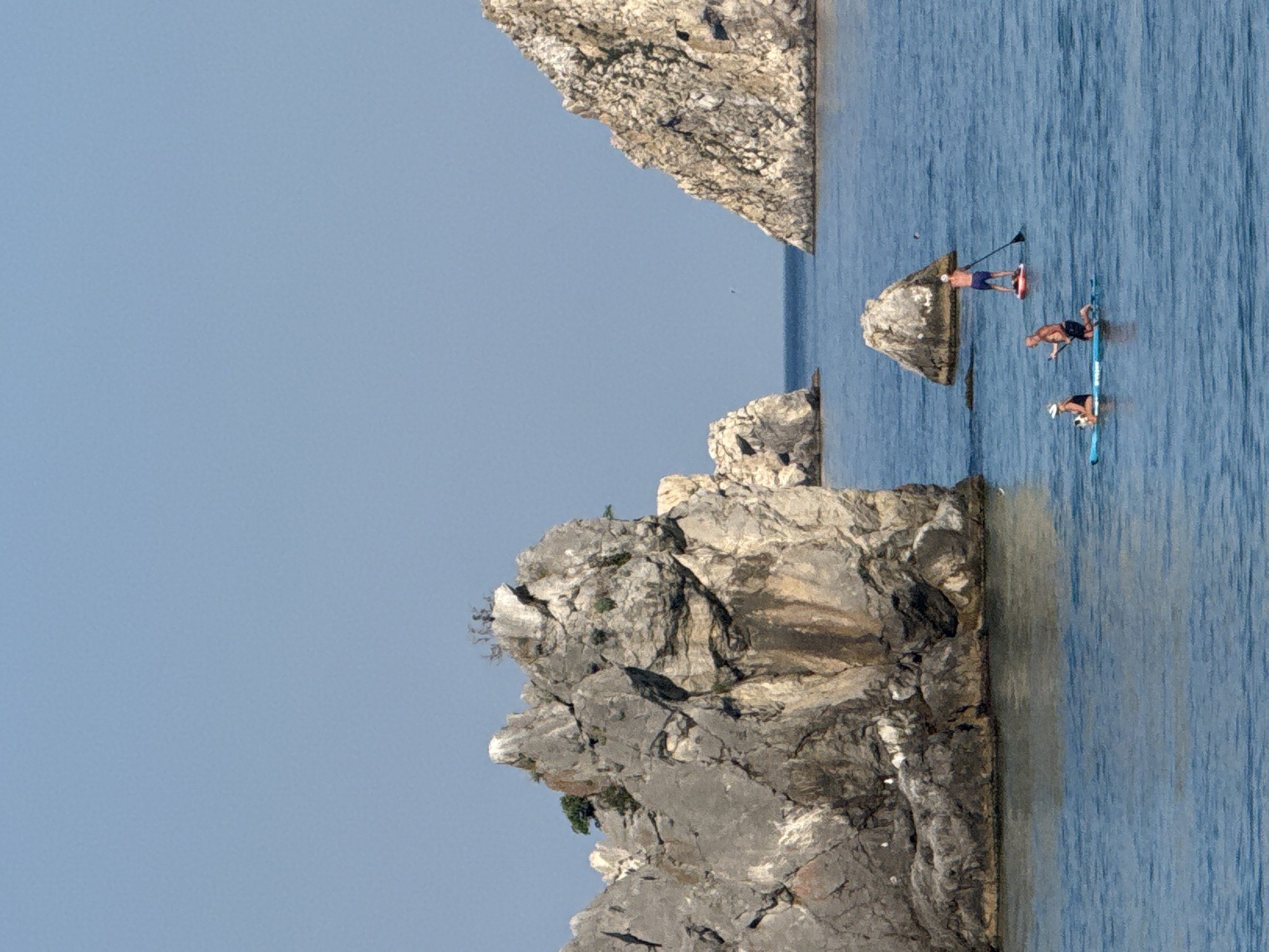 In the image, three people are engaged in paddleboarding on a clear blue body of water, with a backdrop of large, rugged rock formations. The sun is shining, casting shadows and illuminating the scene. One paddleboarder, a male with a white cap and dark shorts, is kneeling and appears to be paddling. Another paddleboarder, a male with a white cap and blue shorts, is standing and also paddling. The third paddleboarder, a female with a white cap and dark shorts, is seated on her board. The rock formations are varied in size and shape, with some appearing to be sea stacks. The sky is a clear, light blue. The environment suggests a coastal or island location.