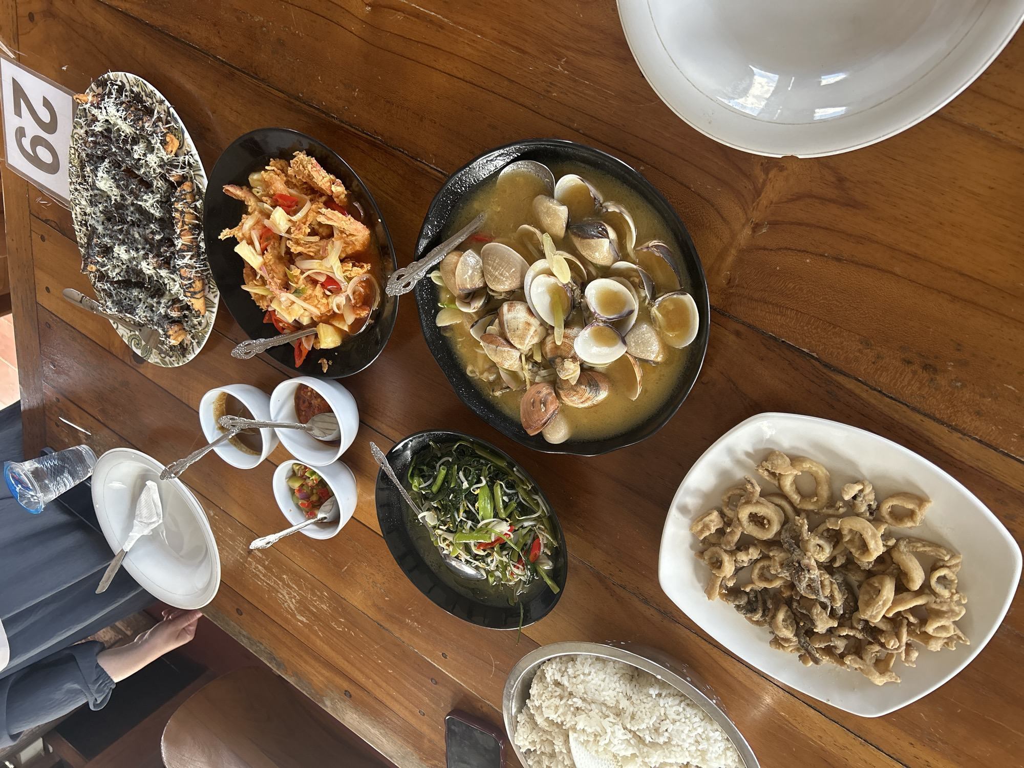 A spread of Indonesian food is displayed on a wooden table. In the foreground, a white oval plate holds a serving of fried calamari rings. To the left, a large metal bowl contains steamed white rice. To the right of the calamari, a black oval bowl is filled with clams in a light yellow broth, garnished with herbs and sliced chilies. Further back, a black oval bowl contains stir-fried water spinach with chili and bean sprouts. A red-chili sauce is served in a small white bowl, alongside another white bowl holding a mixed vegetable relish. A white plate, possibly used or waiting to be served, is positioned at the far right. In the background, a rectangular platter is covered with fried bananas drizzled with chocolate sauce and grated cheese. A table number sign displaying "29" is propped up behind the platter. A person's arm, wearing a dark blue long-sleeved shirt, is visible on the left, holding a white plate. A partially filled plastic cup with a blue lid sits on the table. The scene is indoors, likely in a restaurant.