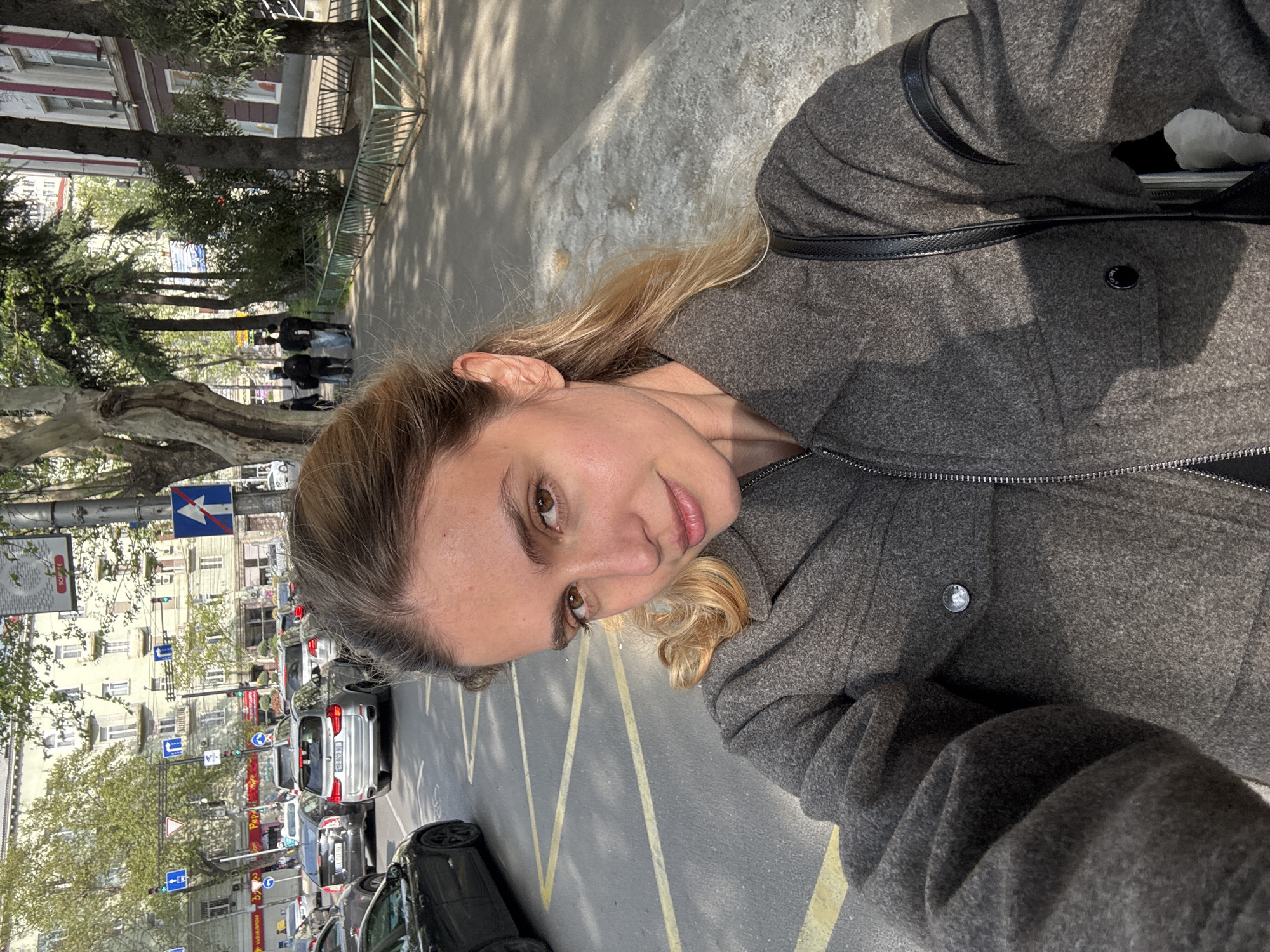 A young woman is captured in a close-up selfie, standing on what appears to be a busy urban street during the daytime. She has medium-length blonde or light brown hair, brown eyes, and fair skin, looking towards the camera with a neutral to slightly pleasant expression. She is wearing a charcoal grey wool-blend jacket with black faux-leather trim along the shoulders and lapel, and a zipper down the front.
Behind her, a bustling city scene unfolds. On the left, several multi-story buildings with various architectural styles, including some with balconies, line the street. Green trees, some with thick trunks, are planted along the sidewalk. A line of cars, both parked and in traffic, is visible on the asphalt road, which features yellow painted lines. Traffic lights and various street signs, including blue circular signs with white arrows indicating mandatory directions, and a blue rectangular sign with a white arrow and a red diagonal line, are present. One car's license plate, visible in traffic, appears to be Georgian with the format "MB-868-HM".
On the right side of the background, more trees with lush green foliage and a metal fence separate the sidewalk from the road. Distant figures of people are seen walking on the sidewalk. The strong, direct sunlight casts clear shadows on the ground and buildings, indicating a bright and sunny day. The overall setting is a vibrant city environment with a mix of old and modern elements.