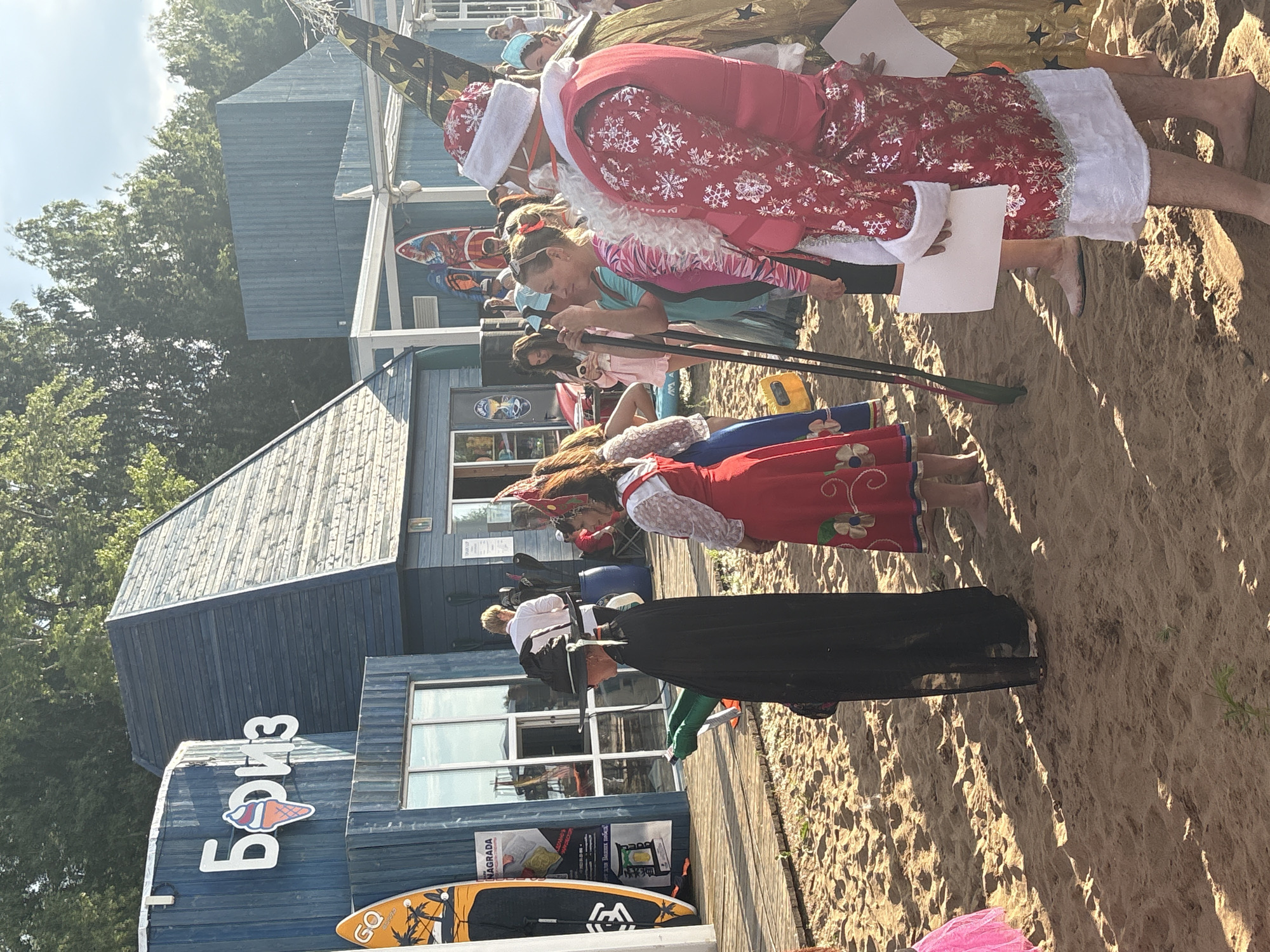 The image shows a sandy area in front of a blue wooden building with the inscription "Борис" and an ice cream cone logo. Trees are visible in the background. Several people are present, some in costumes. In the foreground, a person in a black witch's costume with a pointed hat and green gloves stands on the sand. To their right, a woman in a red dress with white lace and a red headdress is visible. Further to the right, a person dressed as Santa Claus in a red coat with snowflakes and a white beard is holding a white piece of paper. Other individuals, some holding oars, are also in the scene. The environment appears to be a beach or waterfront area, likely in Chaikovsky, Russia, given the presence of the building and the potential context of a recreational area.